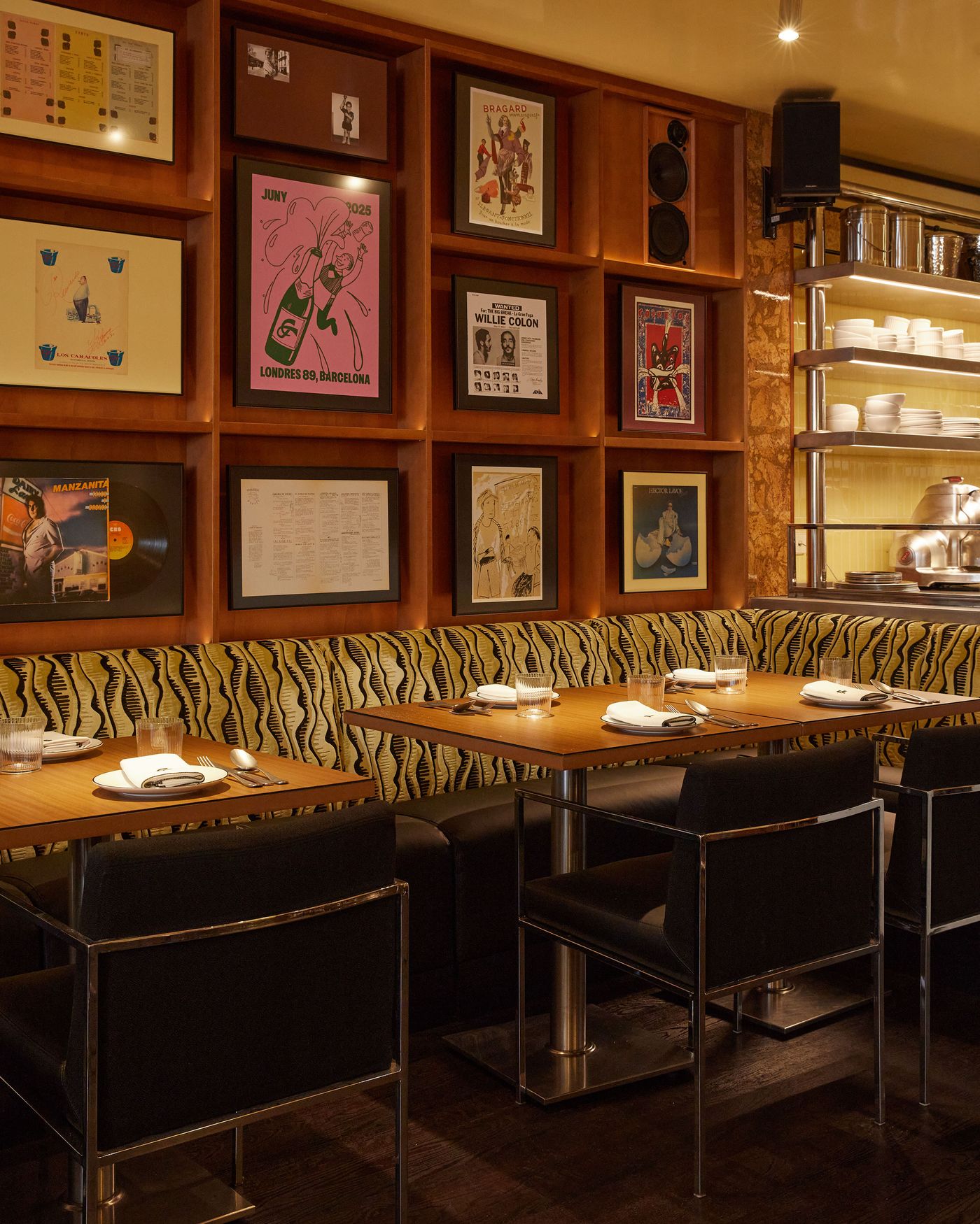 A comprehensive view of a vibrant eclectic dining space. A lively, black and yellow patterned bench seating runs along a wall with a grid of wooden shelves filled with framed art and records. The warm lighting creates an inviting atmosphere, while stainless steel kitchen fixtures are visible in the background. A comprehensive view of a vibrant eclectic dining space. A lively, black and yellow patterned bench seating runs along a wall with a grid of wooden shelves filled with framed art and records. The warm lighting creates an inviting atmosphere, while stainless steel kitchen fixtures are visible in the background.