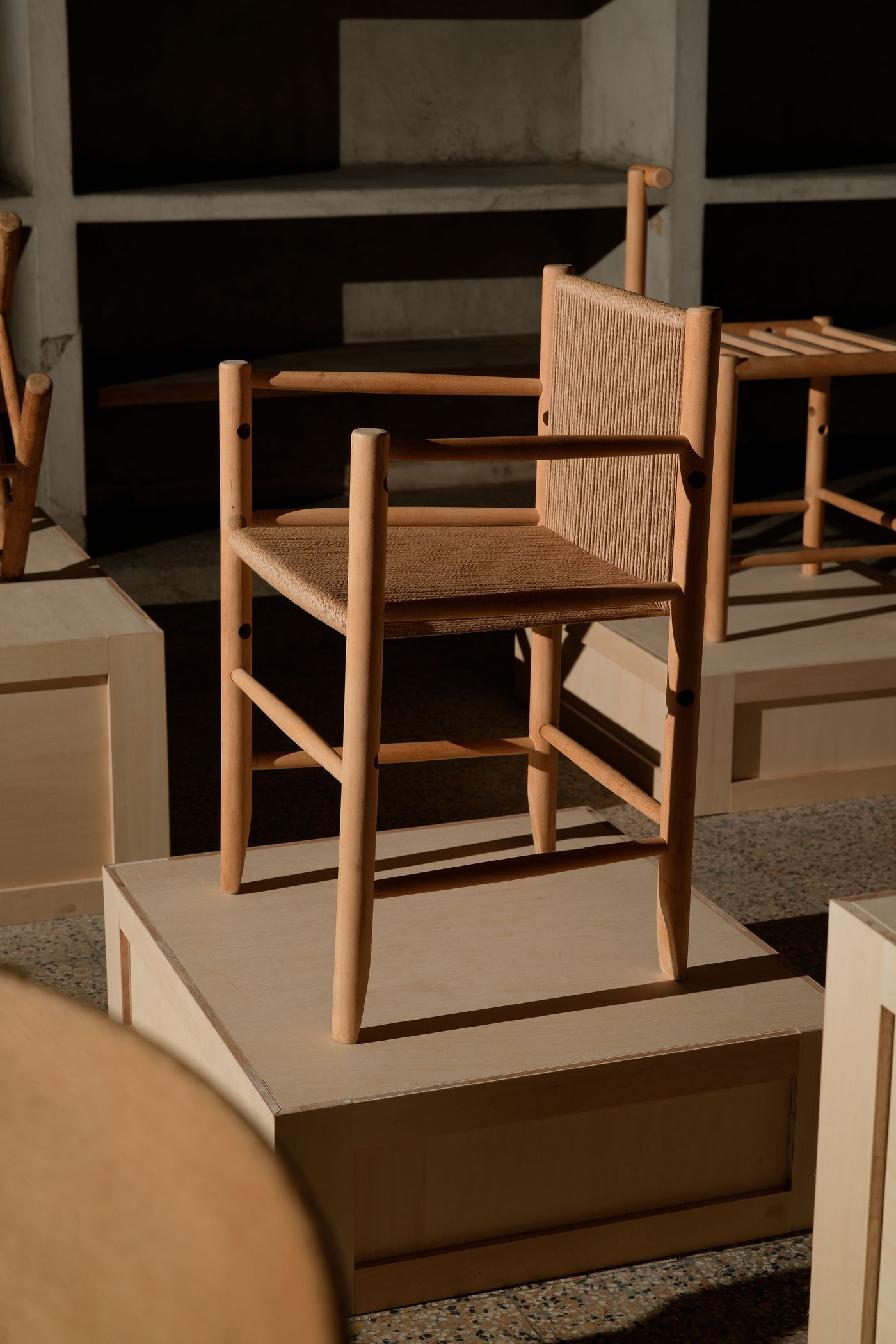 A stylish wooden chair with a woven seat, displayed on a light-colored pedestal amidst other furniture pieces and soft shadows. A stylish wooden chair with a woven seat, displayed on a light-colored pedestal amidst other furniture pieces and soft shadows.