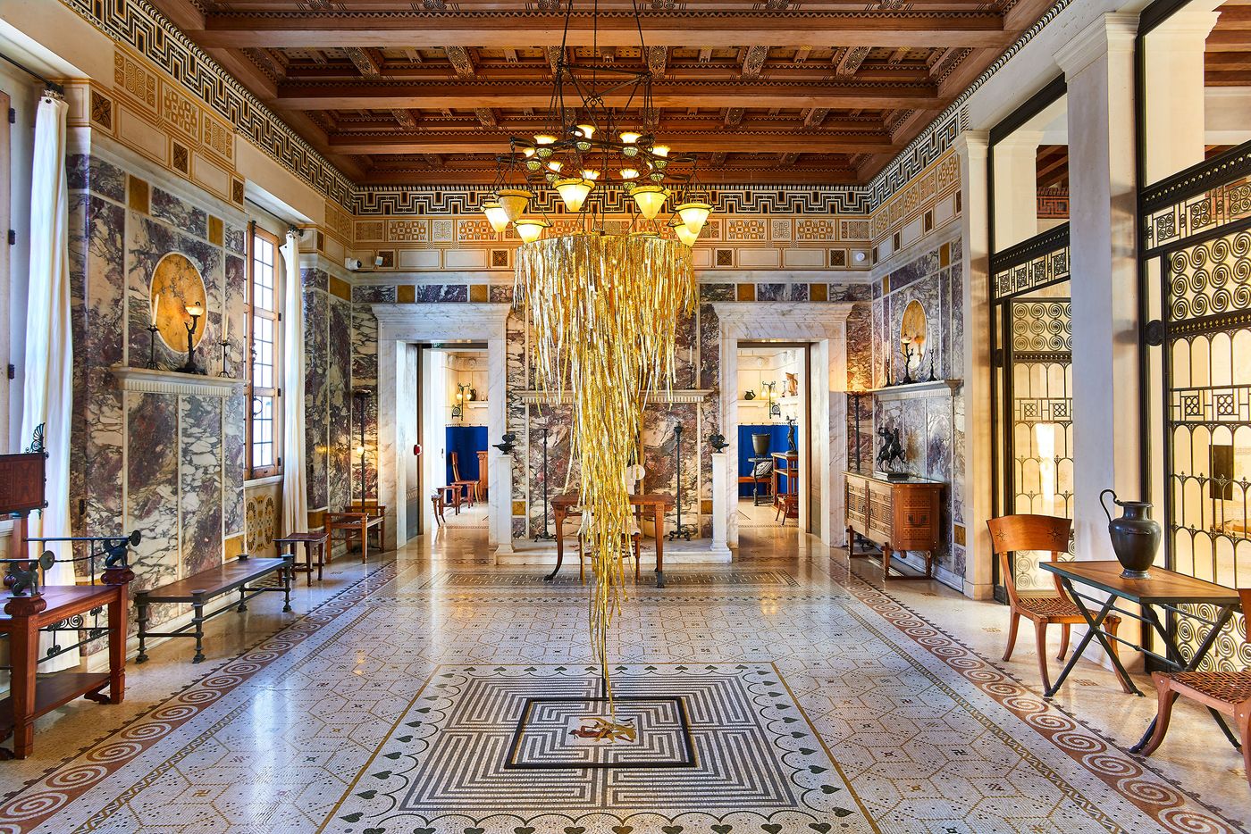 An opulent, symmetrical interior hall with multi-tonal marble walls and a patterned mosaic floor. A contemporary, suspended art installation by Gabriel Leger, featuring a dense cascade of golden, metallic strips, hangs in the center. The piece acts as a modern, glittering focal point within the traditionally styled space, illuminated by two ornate chandeliers. An opulent, symmetrical interior hall with multi-tonal marble walls and a patterned mosaic floor. A contemporary, suspended art installation by Gabriel Leger, featuring a dense cascade of golden, metallic strips, hangs in the center. The piece acts as a modern, glittering focal point within the traditionally styled space, illuminated by two ornate chandeliers.