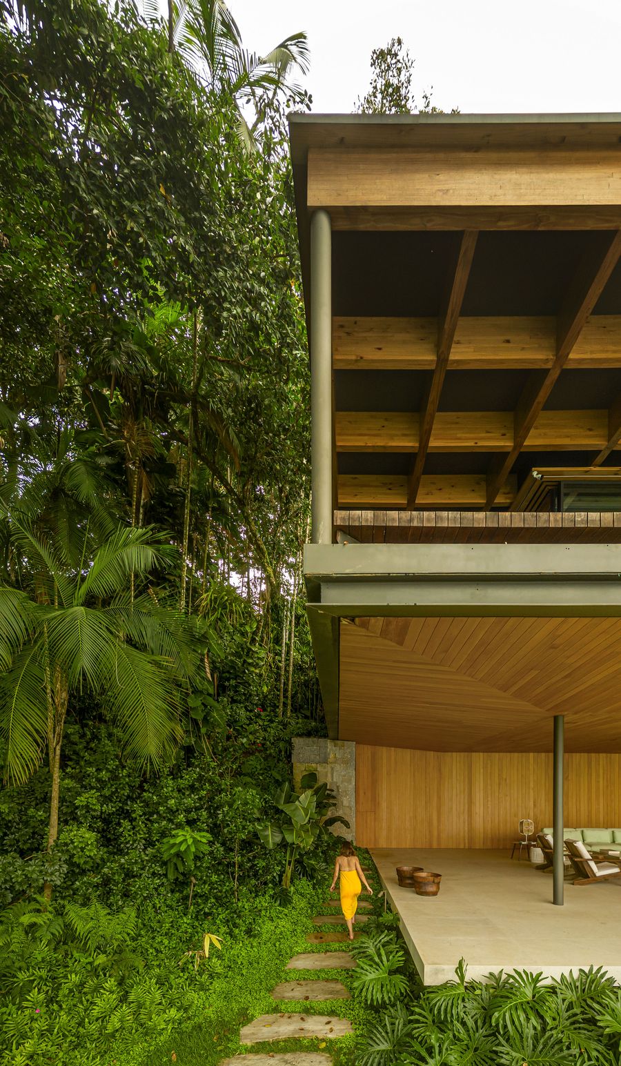 A person in a bright yellow dress descends a natural stone path leading to a sheltered outdoor living area beneath the cantilevered wooden structure. The vibrant tropical foliage cascades around the architectural elements, highlighting the seamless indoor-outdoor transition and the rich material palette of timber and stone. A person in a bright yellow dress descends a natural stone path leading to a sheltered outdoor living area beneath the cantilevered wooden structure. The vibrant tropical foliage cascades around the architectural elements, highlighting the seamless indoor-outdoor transition and the rich material palette of timber and stone.