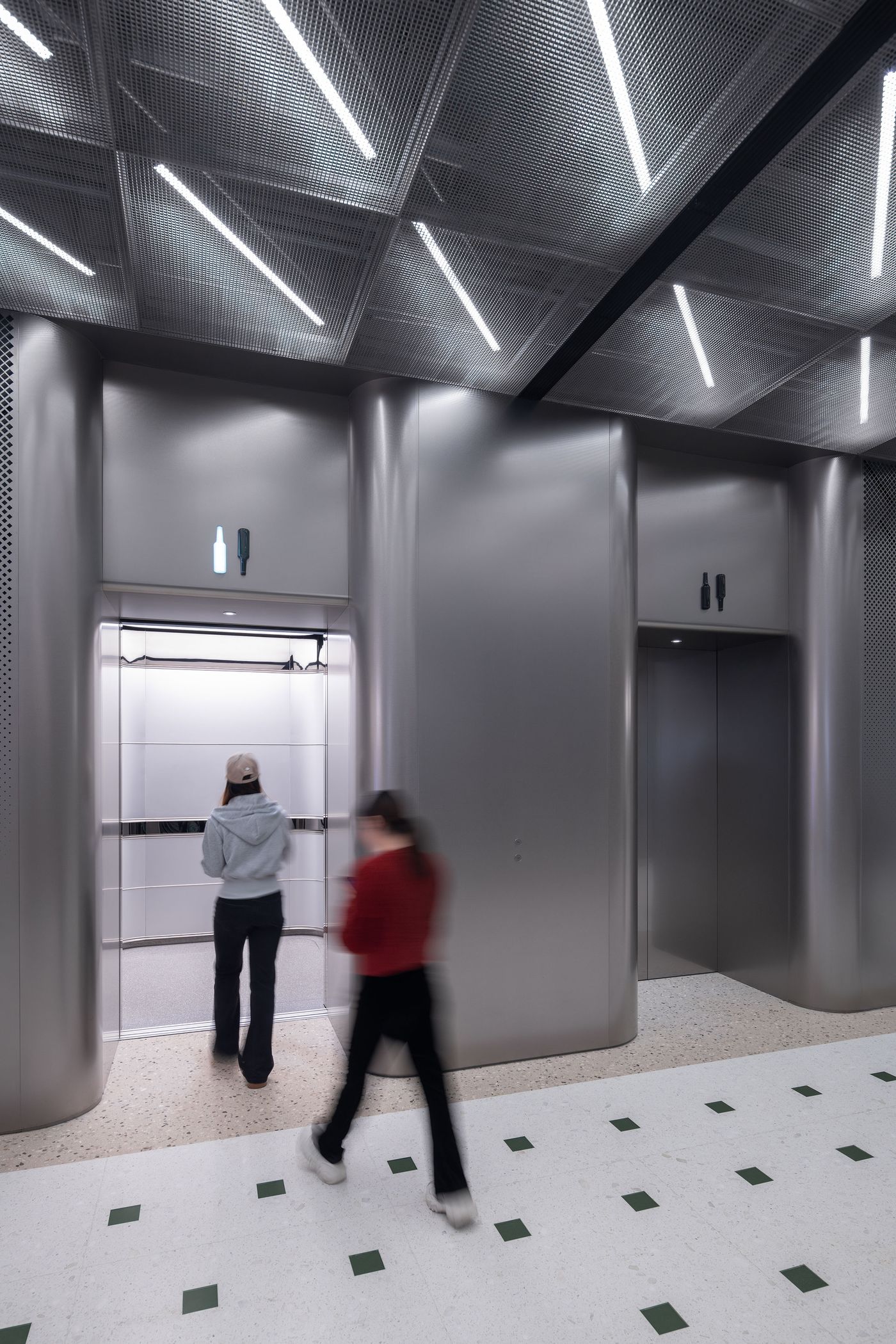 A person in a gray hoodie approaches an elevator, while another walks by, in a modern building with metallic finishes and bright lighting.