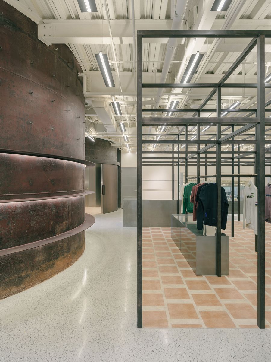 The industrial interior seamlessly blends retail and architectural elements. Towering, curved walls of oxidized steel create a dramatic backdrop, lit from within, contrasting with a lighter retail area defined by a grid of dark metal framing. Garments are displayed on sleek, silver-toned minimalist counters, all beneath an exposed ceiling of white beams and linear lighting. The industrial interior seamlessly blends retail and architectural elements. Towering, curved walls of oxidized steel create a dramatic backdrop, lit from within, contrasting with a lighter retail area defined by a grid of dark metal framing. Garments are displayed on sleek, silver-toned minimalist counters, all beneath an exposed ceiling of white beams and linear lighting.