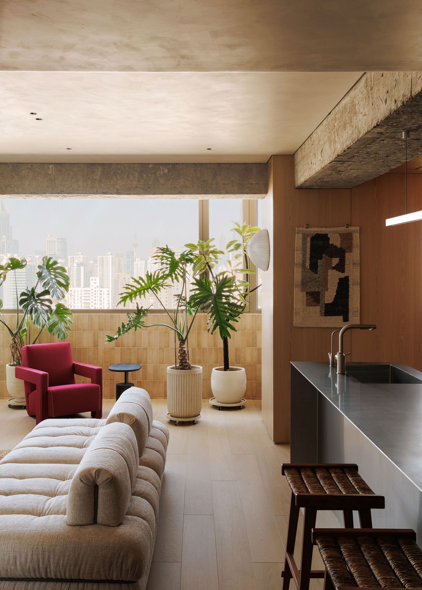 A sophisticated urban living space with panoramic city views, featuring a harmonious blend of industrial and natural elements. Exposed concrete beams contrast with warm wood paneling and a unique ribbed tiled wall beneath the window. A plush cream modular sofa and a bold red armchair offer comfortable seating, while large potted plants introduce a refreshing biophilic touch. A sophisticated urban living space with panoramic city views, featuring a harmonious blend of industrial and natural elements. Exposed concrete beams contrast with warm wood paneling and a unique ribbed tiled wall beneath the window. A plush cream modular sofa and a bold red armchair offer comfortable seating, while large potted plants introduce a refreshing biophilic touch.