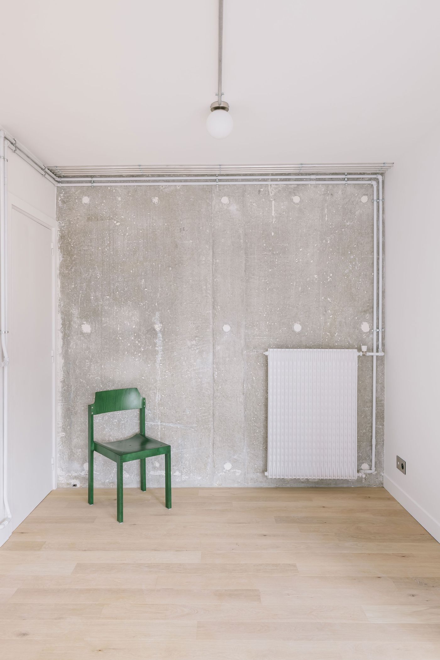 Empty minimalist room with light wooden floor, concrete wall, a green chair, and a white radiator under overhead lighting.