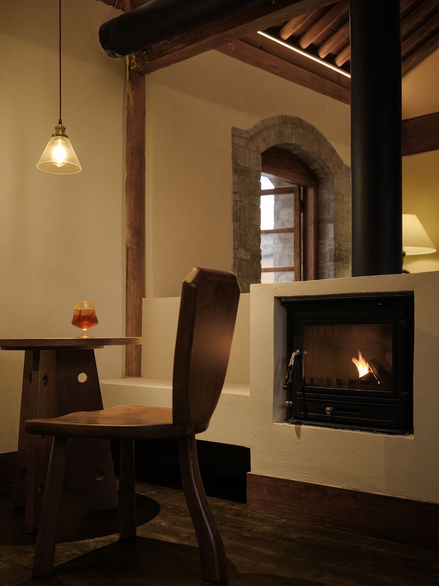 Cozy interior featuring a wooden chair and table, a glowing fireplace, and a drink, illuminated by a pendant light.