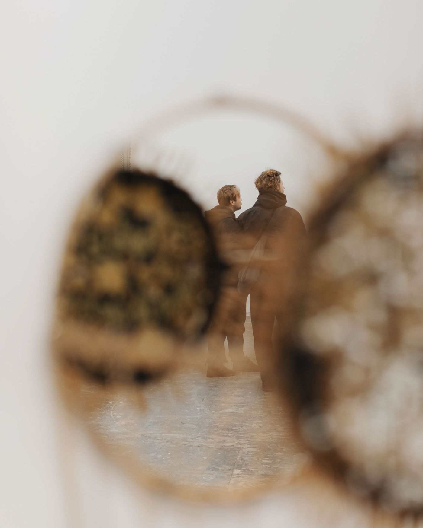 Two figures stand in a gallery, viewed through a circular mirror, surrounded by textures and patterns, part of an artwork on display.