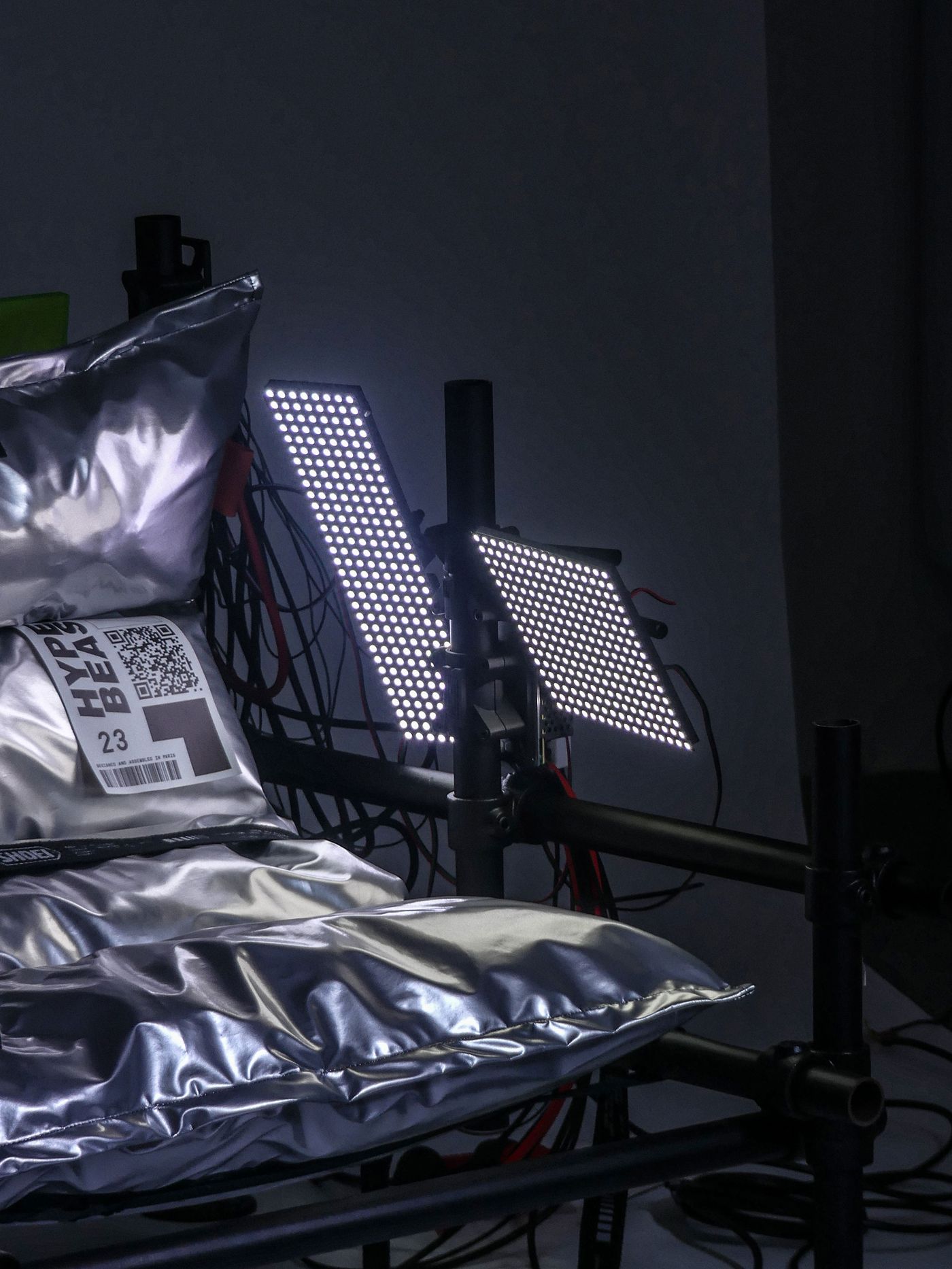 Close-up of a metallic pillow with a barcode, illuminated by two modern LED lights, set against a dark background with cables.