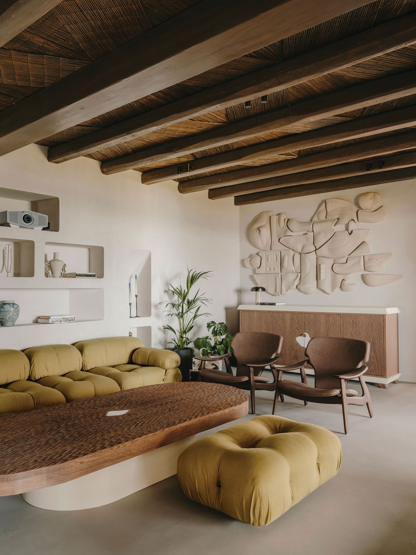 A modern-meets-vernacular living room featuring a curved mustard-coloured sofa, wooden coffee table, two chairs, a textured wall art piece and wooden ceiling meams.