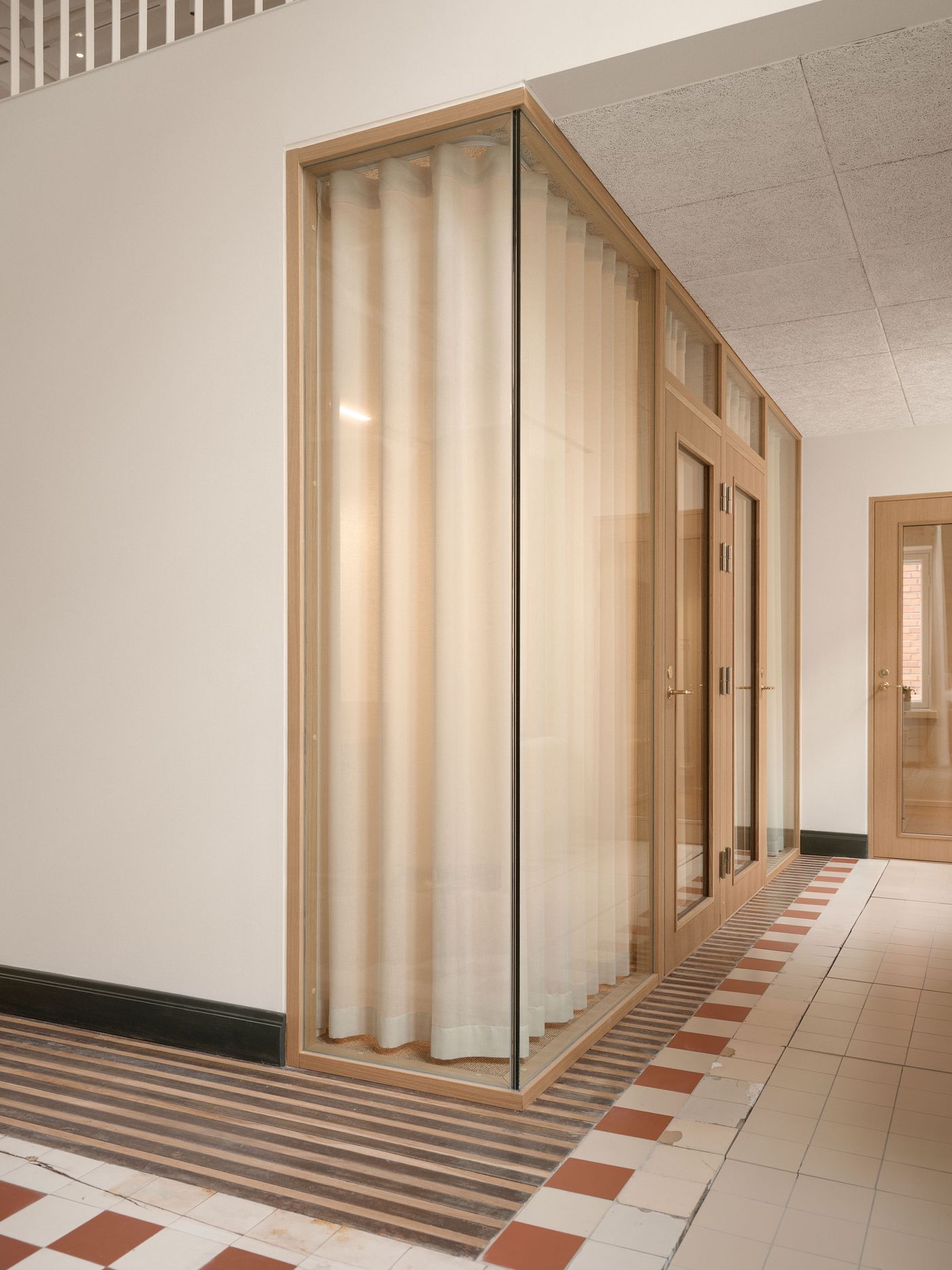 A glass-enclosed office space with sheer curtains provides a private yet transparent work environment within a larger open-plan area. The wooden door frames and the continuation of the distinctive checkerboard flooring emphasize the thoughtful design details.
