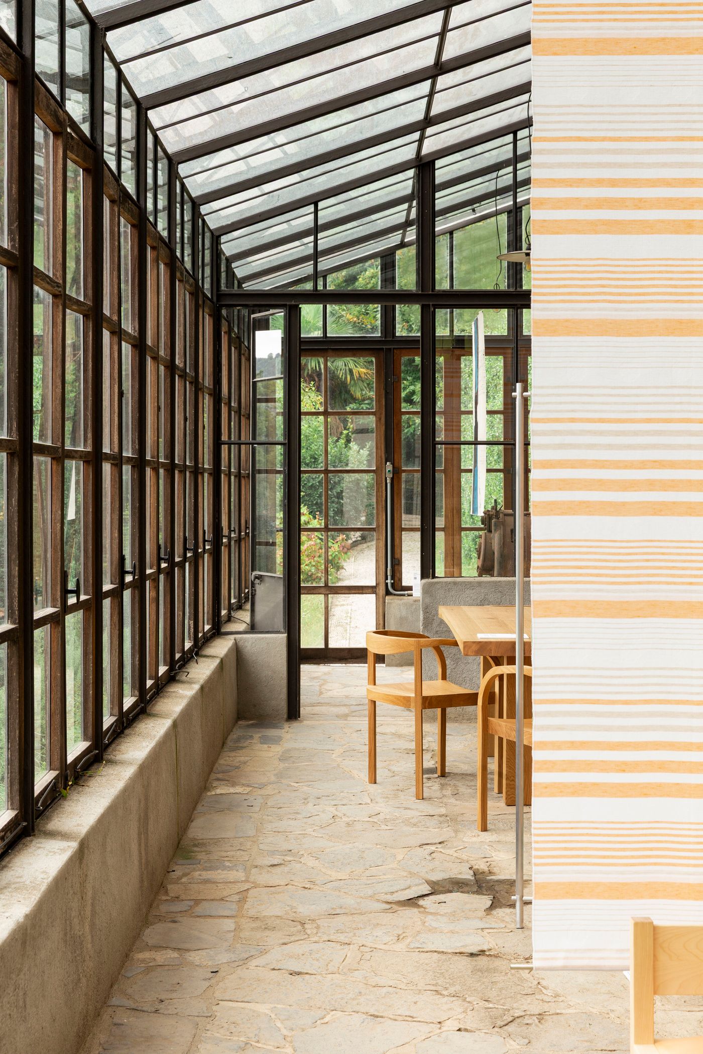 This image captures a bright conservatory or sunroom, with a stone floor and walls of glass panes framed by dark metal. Natural light streams in, illuminating a simple wooden table and chairs. Through the glass, a verdant garden scene is visible, creating a biophilic connection between the interior and the lush exterior. The clean lines of the architecture and furniture offer a serene, minimalist aesthetic, enhanced by the surrounding greenery.