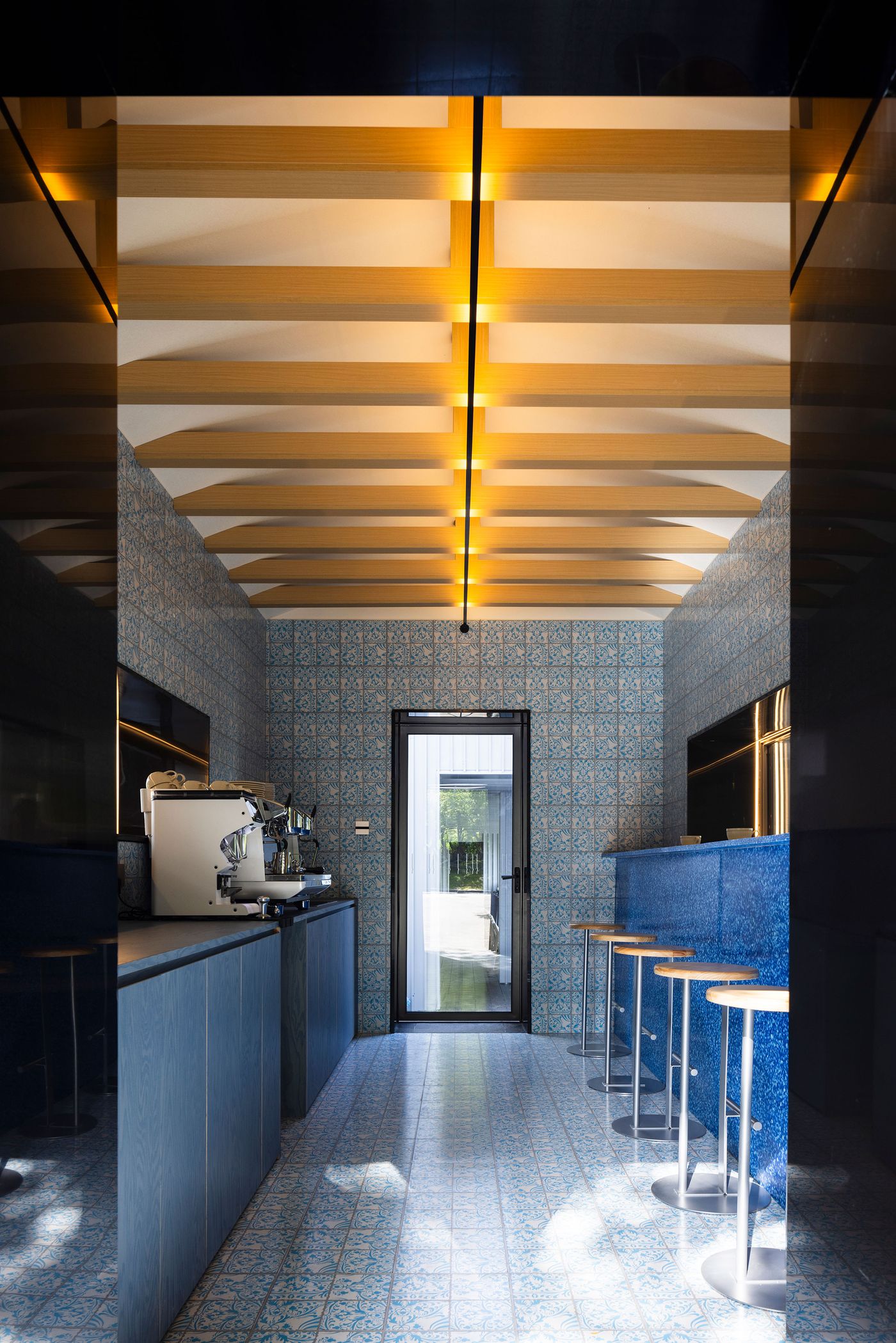 An elongated view into a distinctive interior space, the cafe at Wutopia Lab's Lake House, characterized by its playful blue and white patterned tiles on the walls and floor, creating a Mediterranean or vintage-inspired aesthetic. A sequence of warm, exposed wooden ceiling beams with a central linear light source creates depth, leading to a framed doorway that offers a glimpse of the lush green exterior. An elongated view into a distinctive interior space, the cafe at Wutopia Lab's Lake House, characterized by its playful blue and white patterned tiles on the walls and floor, creating a Mediterranean or vintage-inspired aesthetic. A sequence of warm, exposed wooden ceiling beams with a central linear light source creates depth, leading to a framed doorway that offers a glimpse of the lush green exterior.