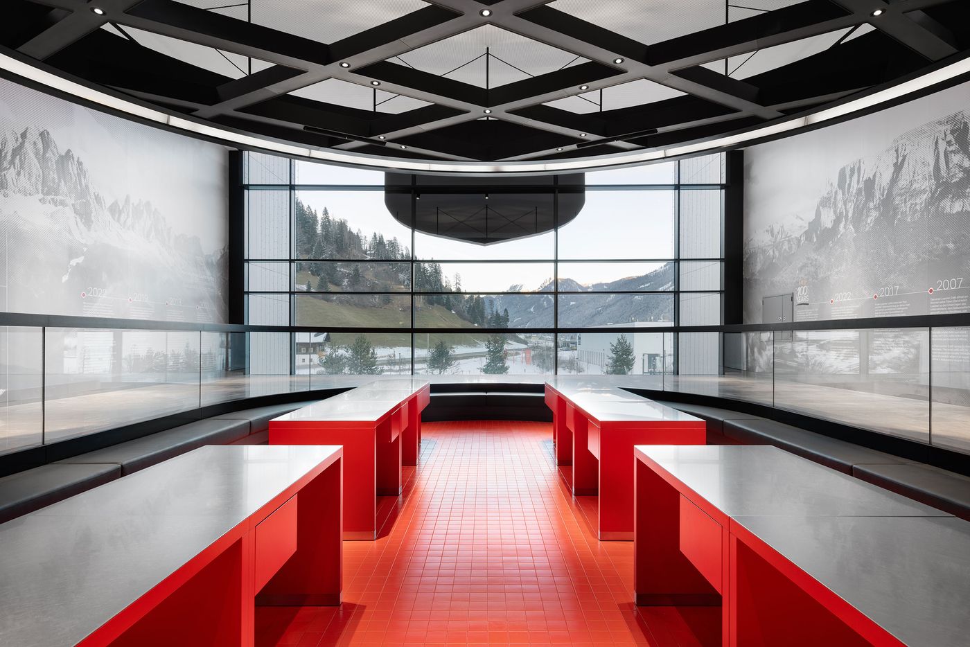 Panoramic alpine vistas stretch across curved exhibition walls beneath a sophisticated grid ceiling system. A sunken red presentation area creates theatrical focus, while the seamless integration of landscape imagery with architectural space demonstrates how interior design can extend natural environment through immersive visual storytelling. Panoramic alpine vistas stretch across curved exhibition walls beneath a sophisticated grid ceiling system. A sunken red presentation area creates theatrical focus, while the seamless integration of landscape imagery with architectural space demonstrates how interior design can extend natural environment through immersive visual storytelling.