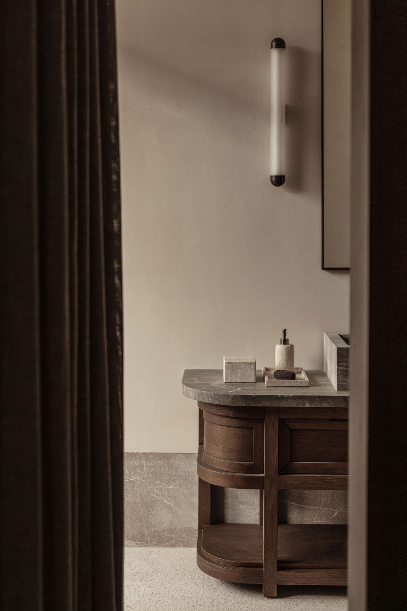 A glimpse into a bathroom reveals a custom-designed dark wood vanity with a curved front and an integrated stone countertop. Contemporary toiletries in muted tones sit elegantly on a wooden tray. The warm, textural wall finish and a modern tube light fixture enhance the room's subtle sophistication, framed by a dark curtain. A glimpse into a bathroom reveals a custom-designed dark wood vanity with a curved front and an integrated stone countertop. Contemporary toiletries in muted tones sit elegantly on a wooden tray. The warm, textural wall finish and a modern tube light fixture enhance the room's subtle sophistication, framed by a dark curtain.