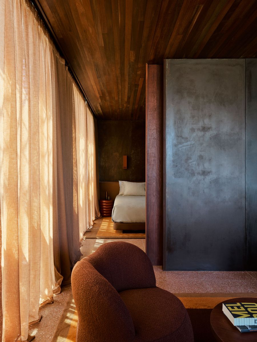 A cozy, modern interior featuring a plush brown chair, sheer curtains, and a bed behind a sleek partition with warm wood accents. A cozy, modern interior featuring a plush brown chair, sheer curtains, and a bed behind a sleek partition with warm wood accents.