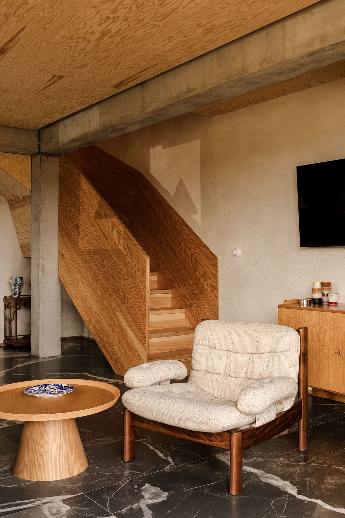 A modern lounge featuring a textured armchair, circular wooden table, and a wooden staircase against a concrete wall and dark marble floor. A modern lounge featuring a textured armchair, circular wooden table, and a wooden staircase against a concrete wall and dark marble floor.