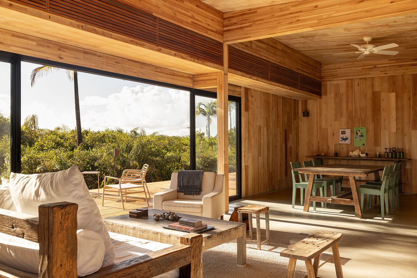 A spacious, light-filled living area features a palette of natural wood and light textiles, offering views of a lush tropical environment through large glass windows. The minimalist furnishings and warm, earthy tones create a serene and organic atmosphere, characteristic of biophilic design.