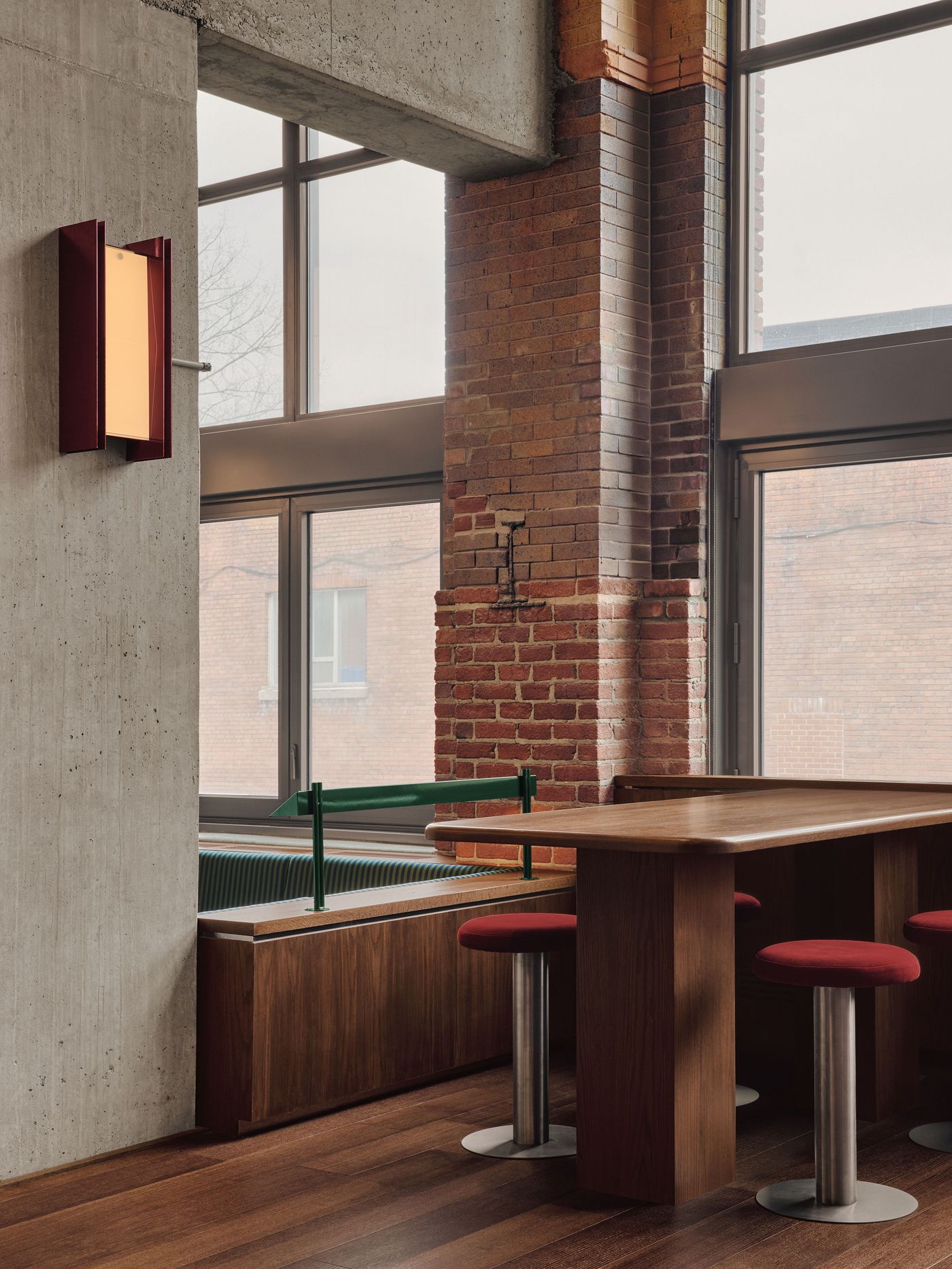 Modern interior featuring a brown wooden table, red stools, and exposed brick walls with large windows illuminating the space.
