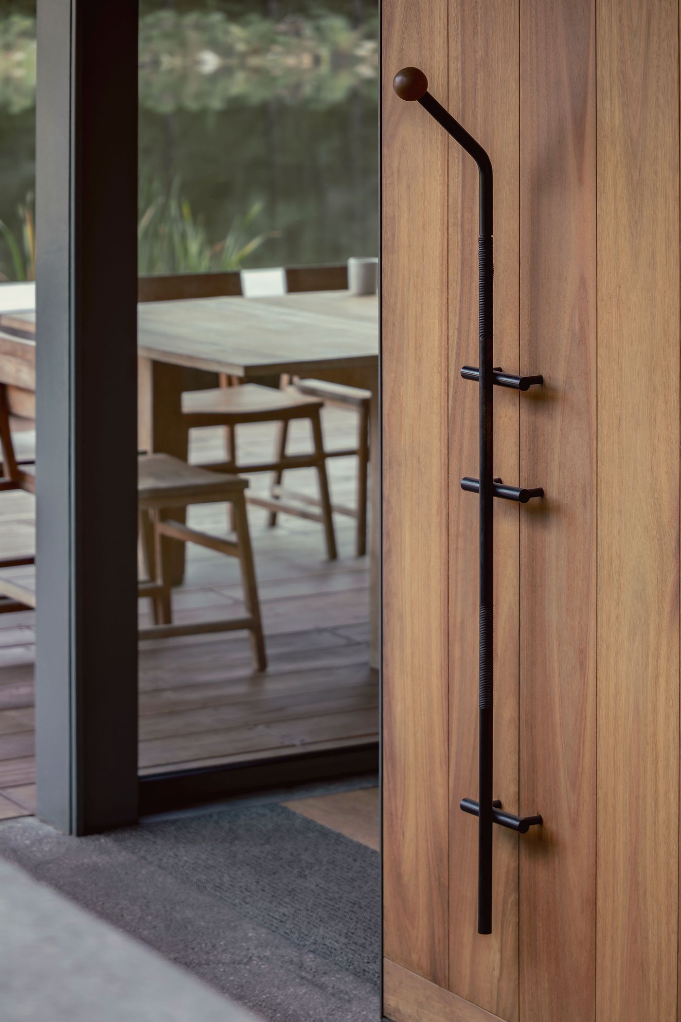 A modern black door handle mounted on wooden wall, with a dining area visible through glass doors in the background.