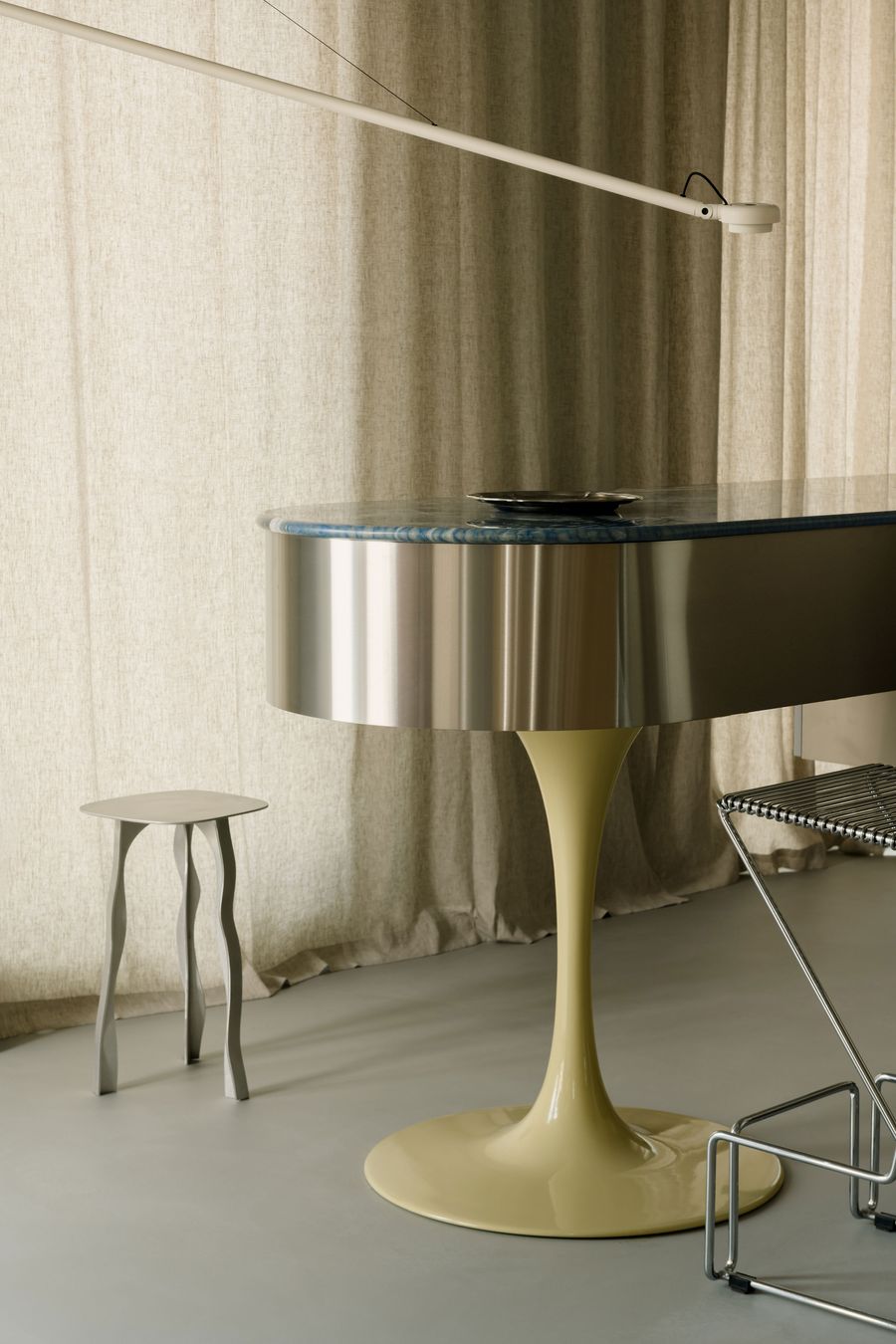 An up-close view of the kitchen island showcases its metallic curvature, topped with a veined stone surface. A slender sculptural stool and retractable arm lamp reinforce the room’s idiosyncratic elegance. An up-close view of the kitchen island showcases its metallic curvature, topped with a veined stone surface. A slender sculptural stool and retractable arm lamp reinforce the room’s idiosyncratic elegance.