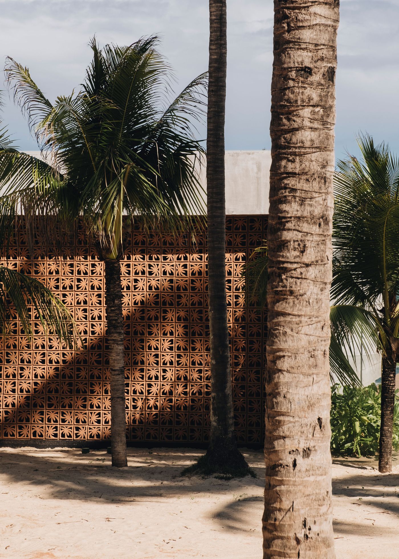 A textured clay wall with decorative patterns stands against swaying palm trees on a sandy beach, under a cloudy sky.