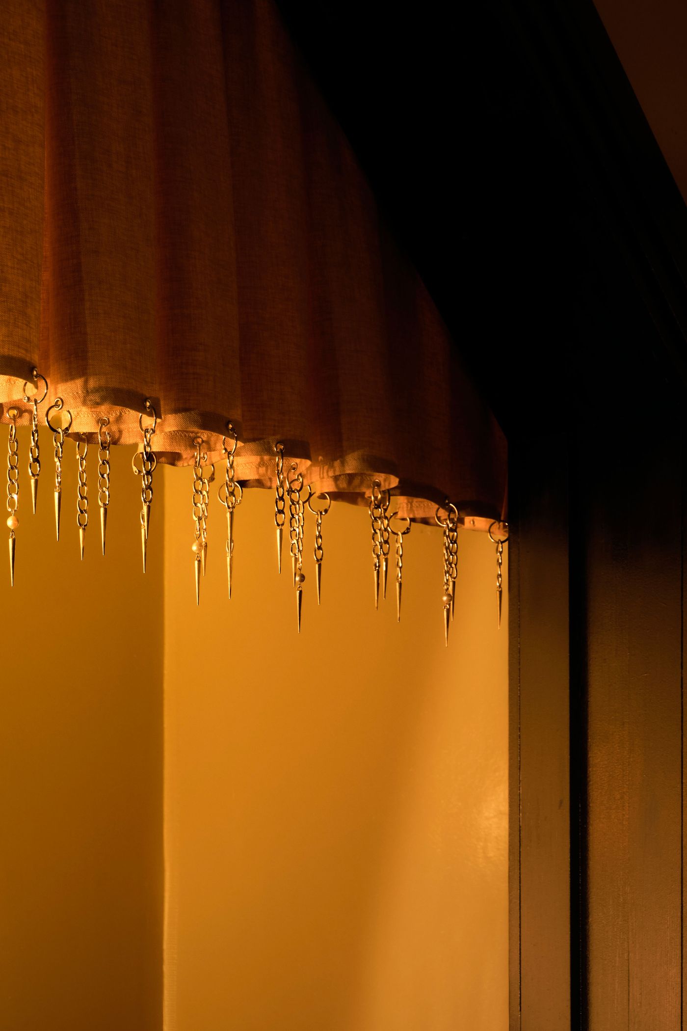 A detailed close-up reveals intricate, chain-linked adornments with pointed, icicle-like drops, suspended from a warm, orange-toned curtain or valance. The dark background creates contrast, highlighting the unique, almost gothic yet elegant detail that contributes to the rich, multi-layered aesthetic of The Roses of Elagabalus.