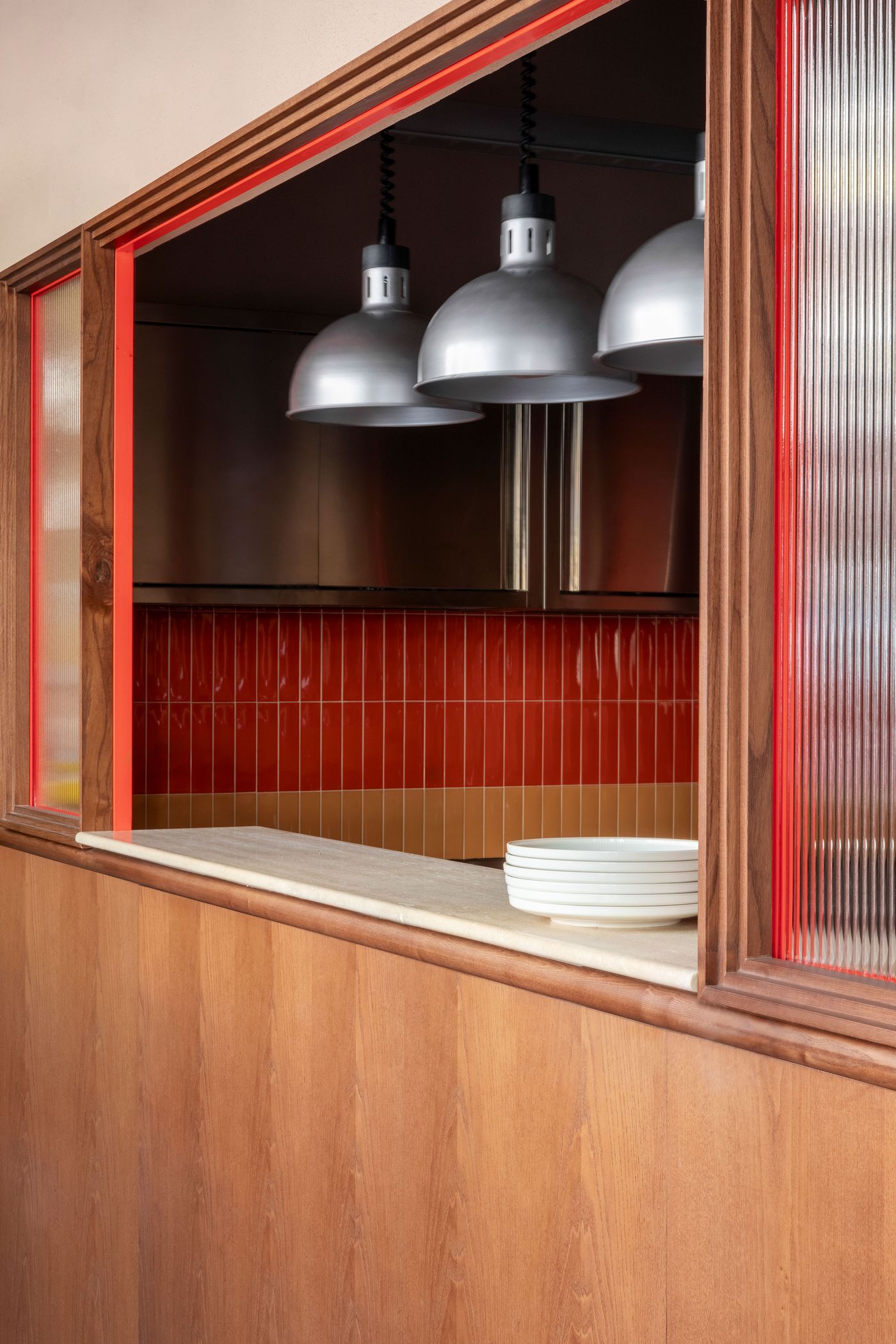 A close-up of Santo Palato's service counter, designed by Naessi Studio, featuring a vibrant two-tone tiled backsplash in red and orange. Silver industrial-style pendant lights hang above a polished marble counter with neatly stacked white bowls. Red-outlined wooden frames with fluted glass add a distinctive, modern touch to the space.