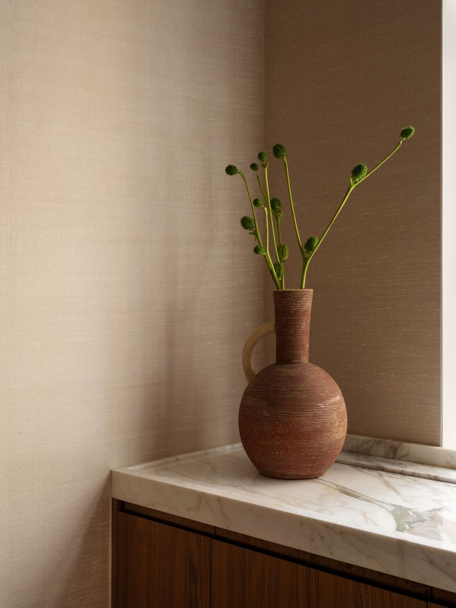 A textured brown vase on a marble countertop, adorned with green, spherical plant stems against a soft beige wall. A textured brown vase on a marble countertop, adorned with green, spherical plant stems against a soft beige wall.