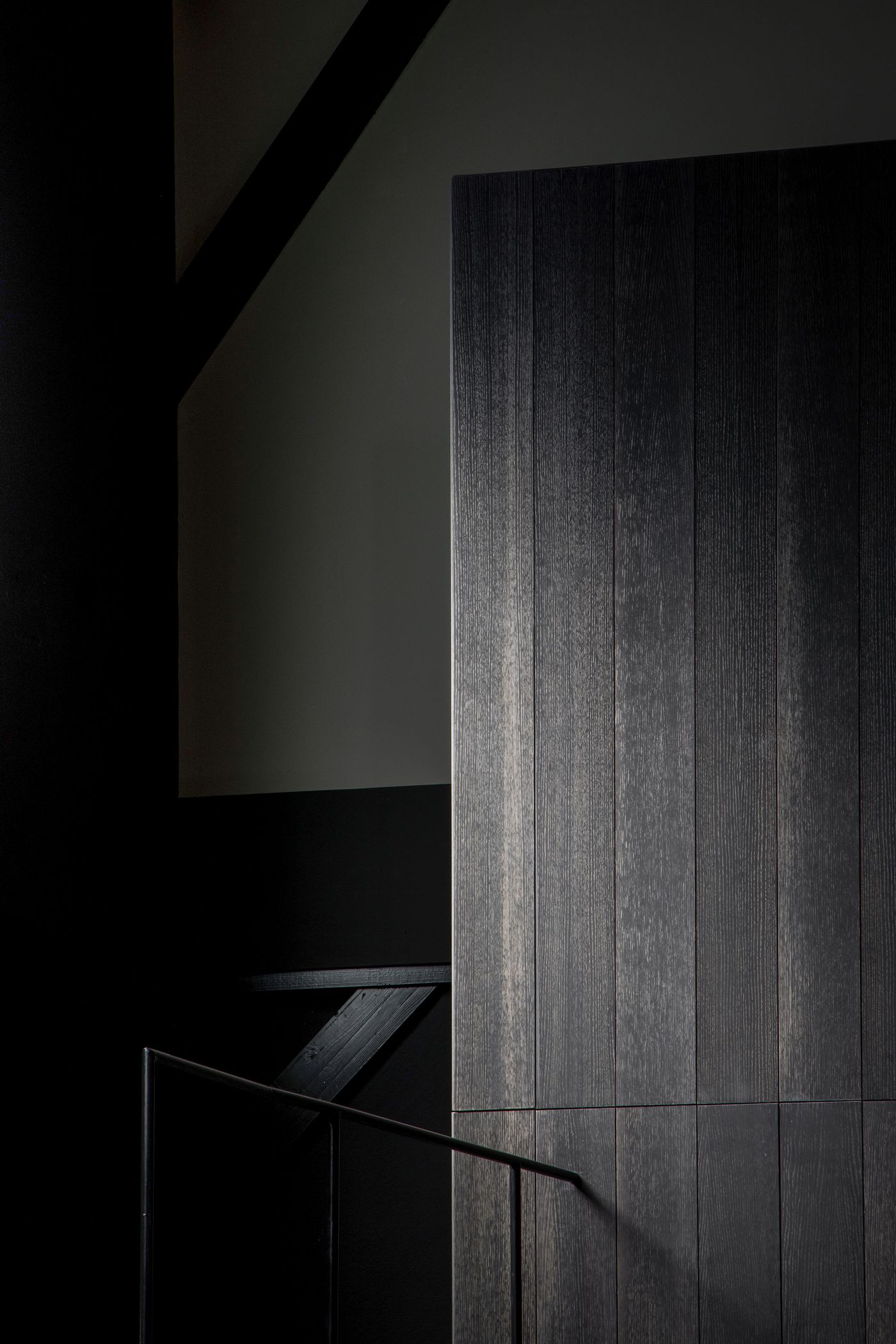 A dark wooden panel with vertical grain texture stands against a minimalist backdrop, with a black railing leading up a staircase.