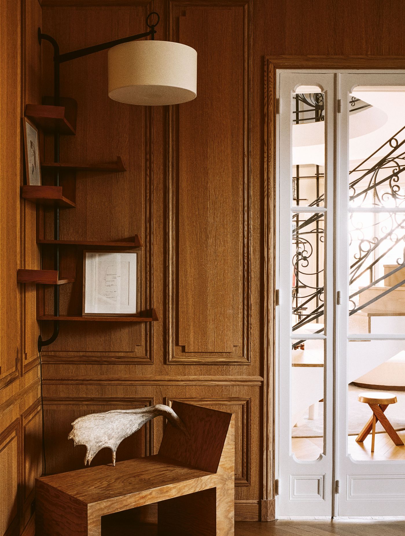 A thoughtfully composed corner of a room, adorned with warm wooden wall paneling. A multi-tiered, suspended wooden shelf unit displays small art pieces, while a cylindrical wall-mounted lamp casts a soft glow. In the foreground, a sculptural chair with a unique fur detail offers an artistic touch. A thoughtfully composed corner of a room, adorned with warm wooden wall paneling. A multi-tiered, suspended wooden shelf unit displays small art pieces, while a cylindrical wall-mounted lamp casts a soft glow. In the foreground, a sculptural chair with a unique fur detail offers an artistic touch.