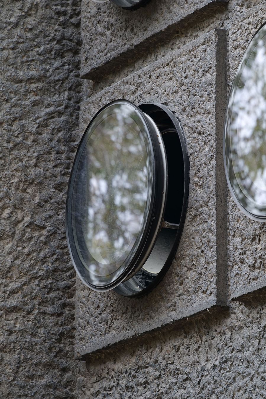 A circular, reflective window set into a concrete-textured gray wall, reflecting shades of green from surrounding trees. A circular, reflective window set into a concrete-textured gray wall, reflecting shades of green from surrounding trees.