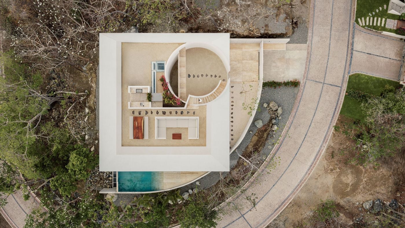 A top-down view of Casa Malonic shows its geometric logic: a crisp white square framing a sand-toned roof terrace, cut by a circular courtyard traced with dark stepping stones. Built-in seating, a timber table and a burst of magenta blooms punctuate the plan, while a curved concrete wall, with the pool tucked along its inner edge, follows the arc of the road. A top-down view of Casa Malonic shows its geometric logic: a crisp white square framing a sand-toned roof terrace, cut by a circular courtyard traced with dark stepping stones. Built-in seating, a timber table and a burst of magenta blooms punctuate the plan, while a curved concrete wall, with the pool tucked along its inner edge, follows the arc of the road.