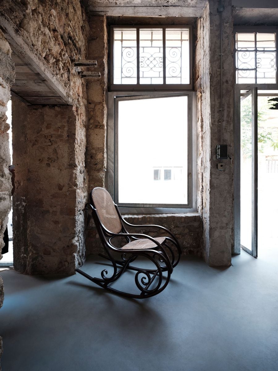 A vintage bentwood-and-cane rocking chair sits beside one of the reopened street-facing openings, surrounded by exposed stone and concrete. Decorative ironwork survives above the new glazing, adding another historical layer, while daylight washes across the seamless grey floor and the chair’s sculptural curved frame. A vintage bentwood-and-cane rocking chair sits beside one of the reopened street-facing openings, surrounded by exposed stone and concrete. Decorative ironwork survives above the new glazing, adding another historical layer, while daylight washes across the seamless grey floor and the chair’s sculptural curved frame.