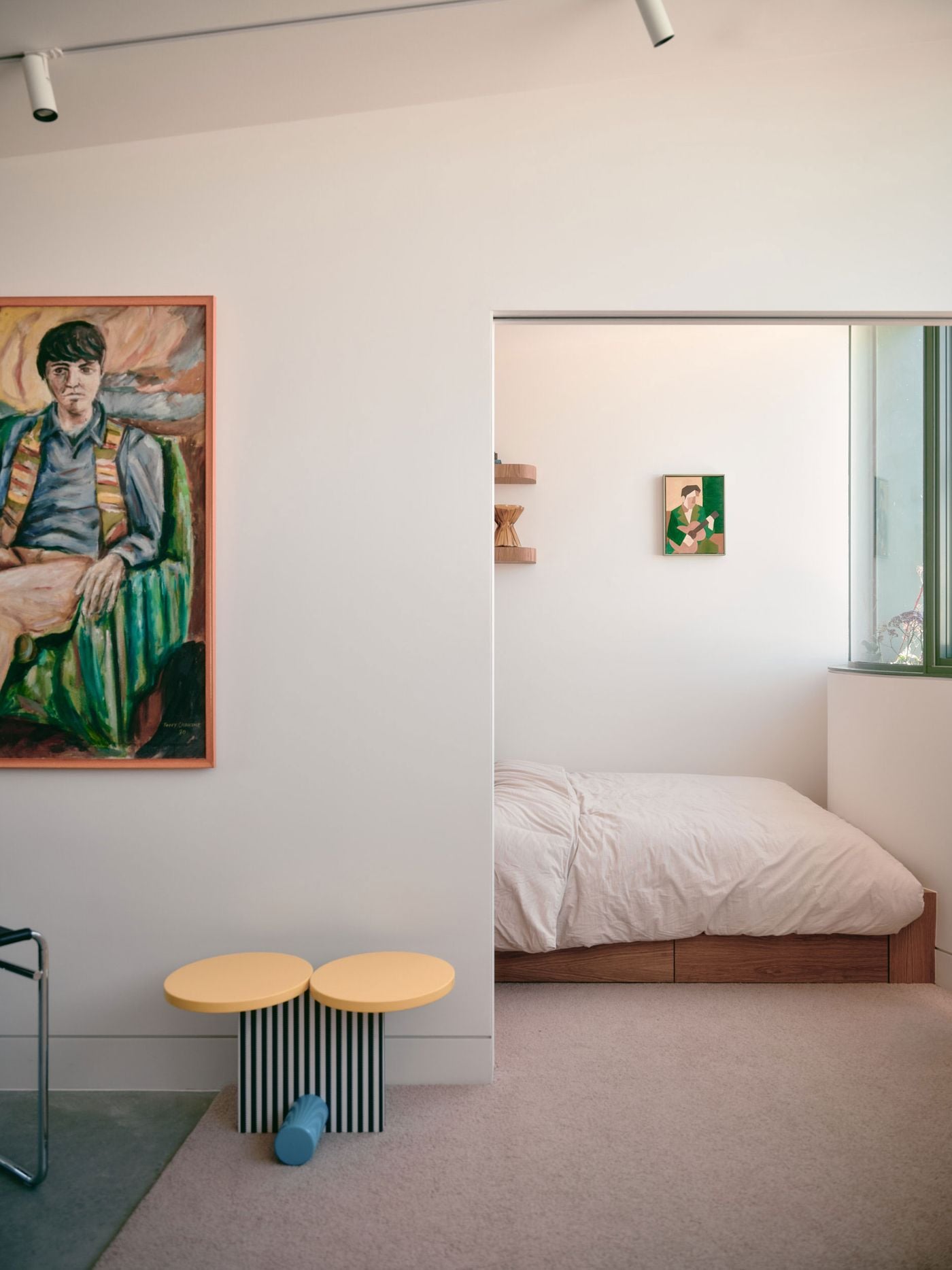 The studio’s living area opens towards a compact bedroom through a broad sliding doorway, maintaining visual continuity across the small plan. A large portrait, striped side table and tubular-steel chair add personality to the restrained setting, while the timber bed and floating shelves introduce warmth and built-in efficiency. The studio’s living area opens towards a compact bedroom through a broad sliding doorway, maintaining visual continuity across the small plan. A large portrait, striped side table and tubular-steel chair add personality to the restrained setting, while the timber bed and floating shelves introduce warmth and built-in efficiency.