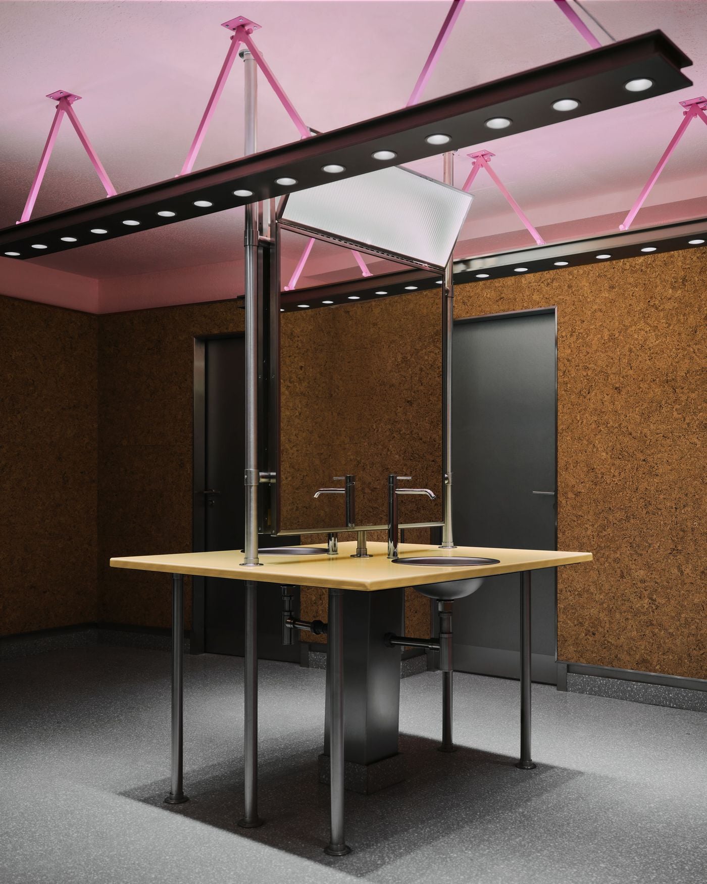 A central wash station in the men’s locker room pairs a butter-yellow countertop with exposed stainless-steel basins and pipework. Cork walls, black doors and suspended linear lights are washed in pink overhead, creating an unexpectedly playful palette within the otherwise utilitarian setting. A central wash station in the men’s locker room pairs a butter-yellow countertop with exposed stainless-steel basins and pipework. Cork walls, black doors and suspended linear lights are washed in pink overhead, creating an unexpectedly playful palette within the otherwise utilitarian setting.