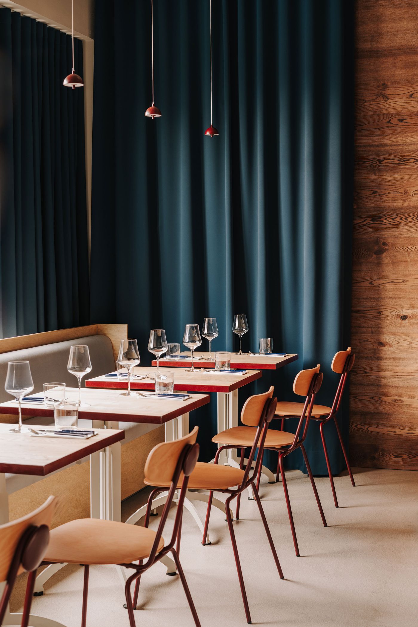 The upstairs dining area at WIN wine bar features a row of timber-topped tables with burgundy-edged surfaces, paired with bentwood chairs supported by slender metal frames. Deep blue floor-to-ceiling curtains and timber wall panelling create a warm, intimate atmosphere, while miniature burgundy pendant lights punctuate the otherwise restrained composition. The upstairs dining area at WIN wine bar features a row of timber-topped tables with burgundy-edged surfaces, paired with bentwood chairs supported by slender metal frames. Deep blue floor-to-ceiling curtains and timber wall panelling create a warm, intimate atmosphere, while miniature burgundy pendant lights punctuate the otherwise restrained composition.