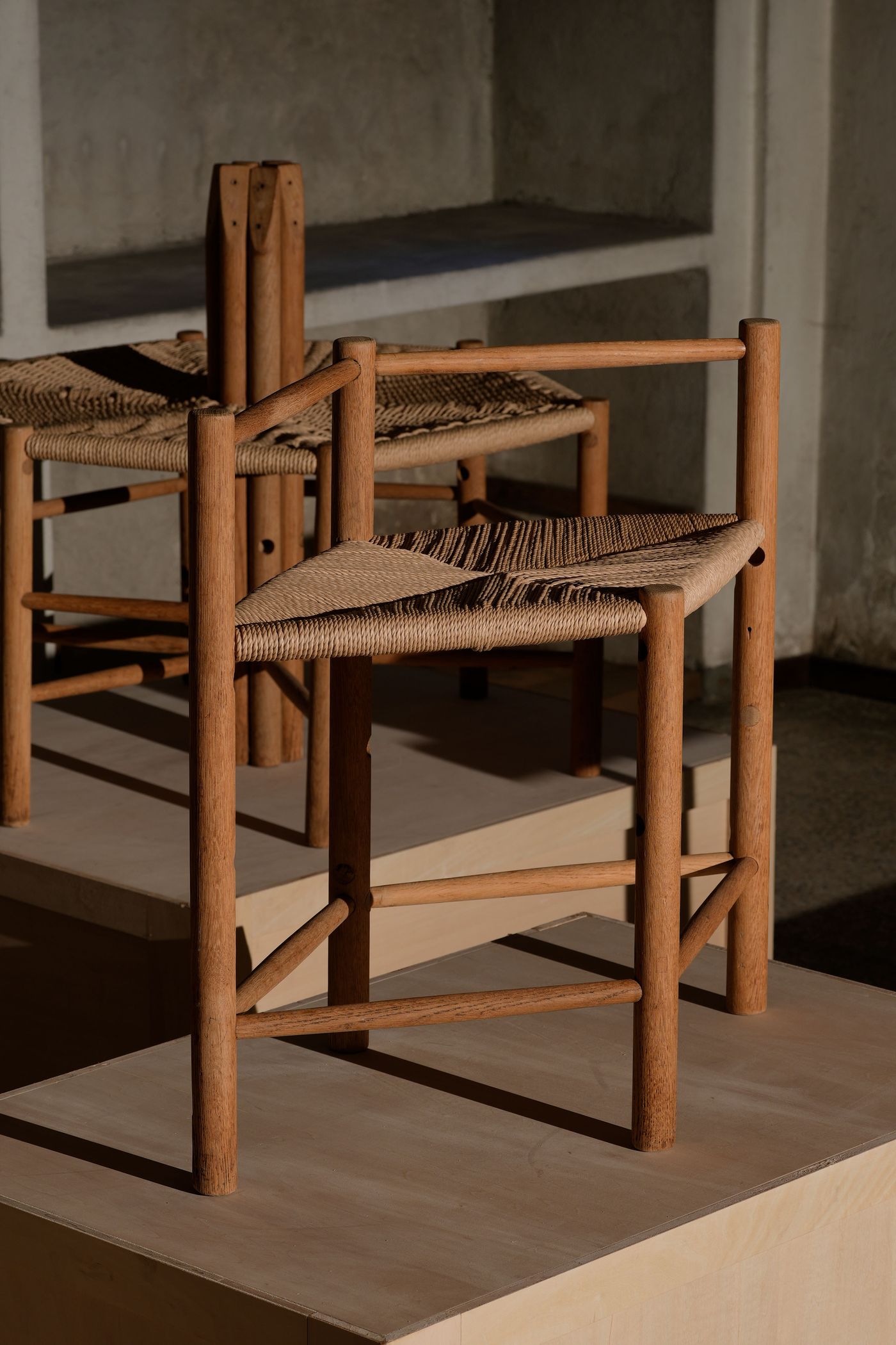 A minimalist wooden chair with a woven seat stands on a wooden platform, showcasing its elegant design against a textured wall backdrop.