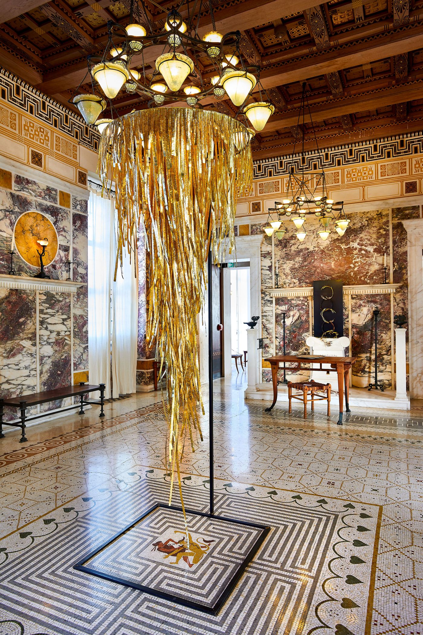 A detail shot of the large, suspended sculpture by Gabriel Leger, a flowing column of golden metallic strips. The piece hangs in the center of a mosaic floor with a bold geometric pattern, framed by a traditional chandelier. This modern, dynamic artwork provides a striking contrast to the classical marble walls and dark wood ceiling.
