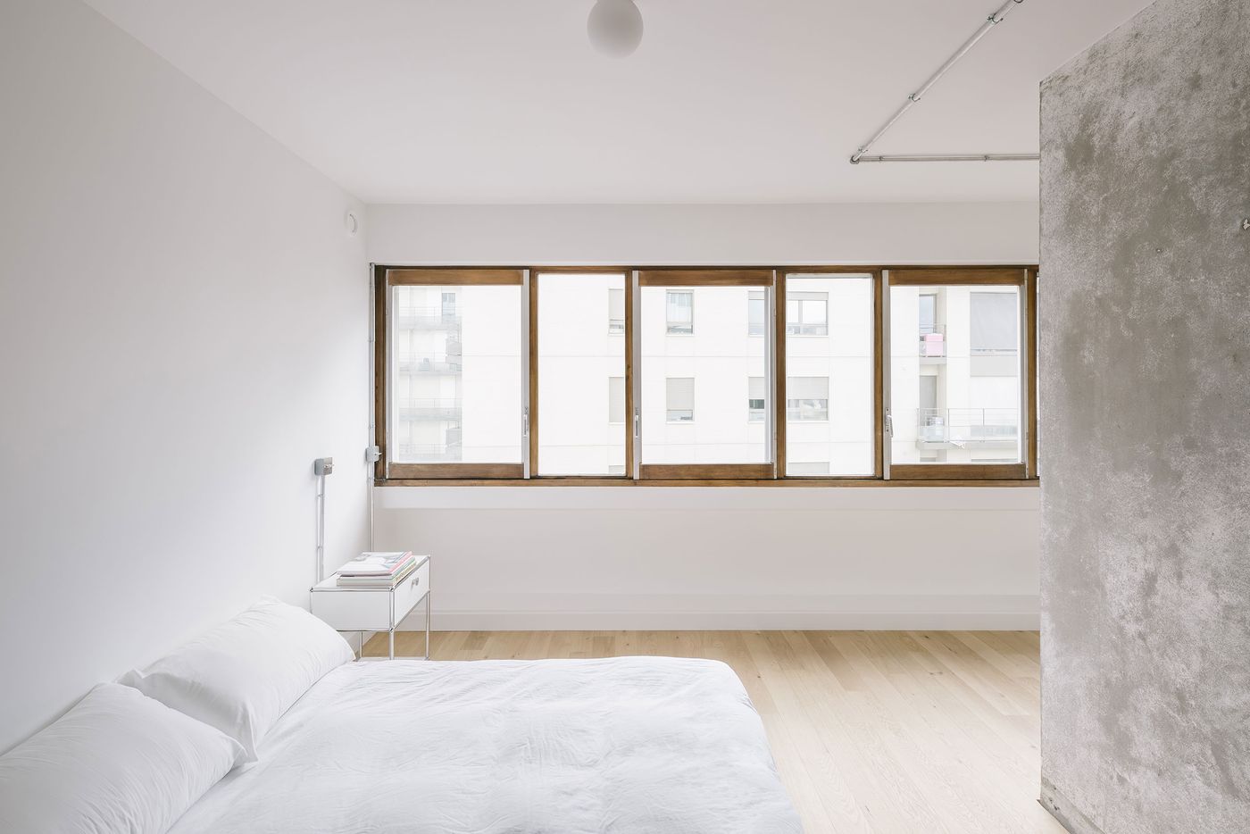 A minimalist bedroom featuring a white bed, modern side table, and large wooden windows overlooking an urban view.