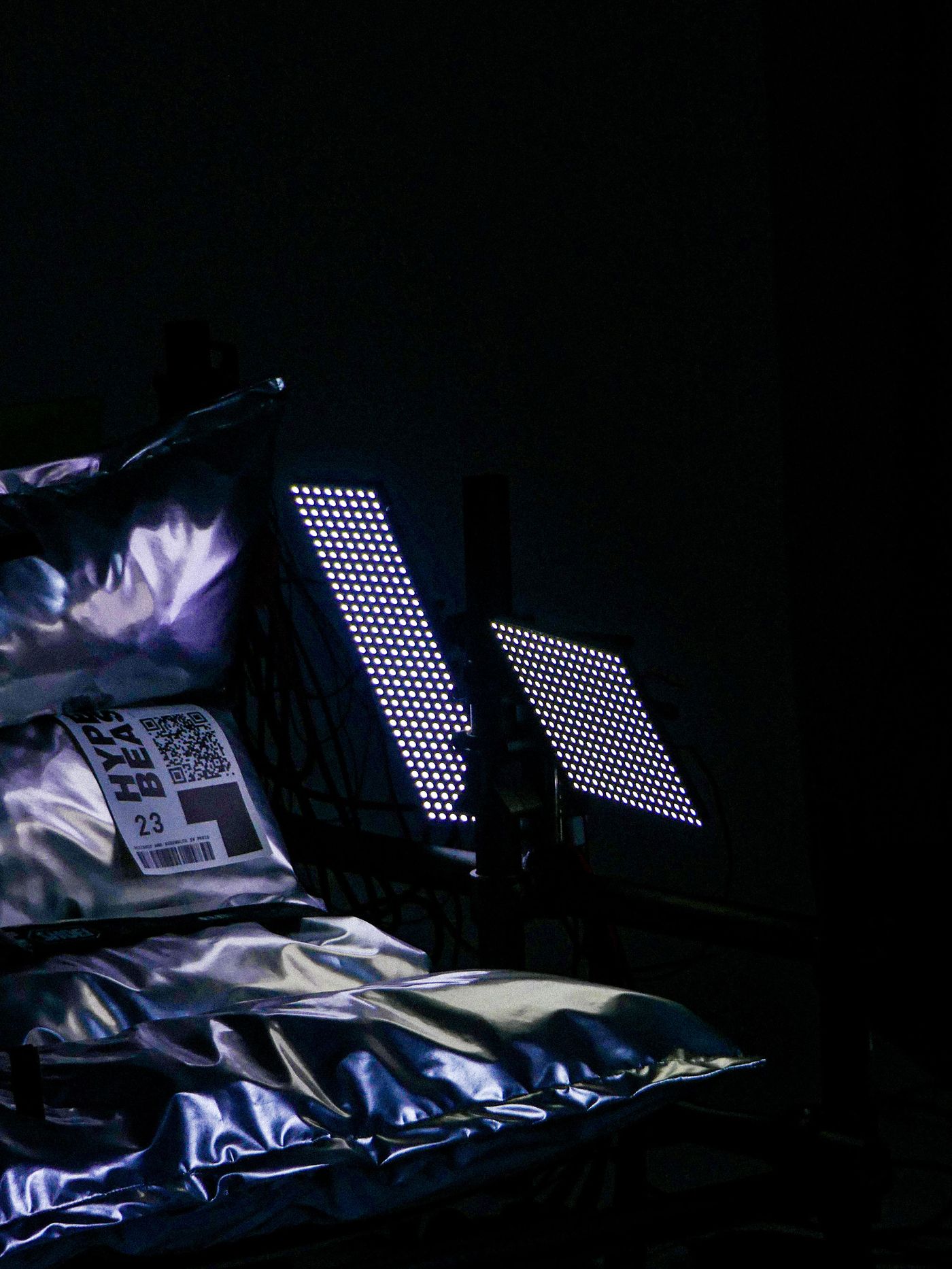 Two illuminated LED panels shine in a dark space beside a shiny, crumpled silver pillow and a package labeled "HYPERBEAT 23."