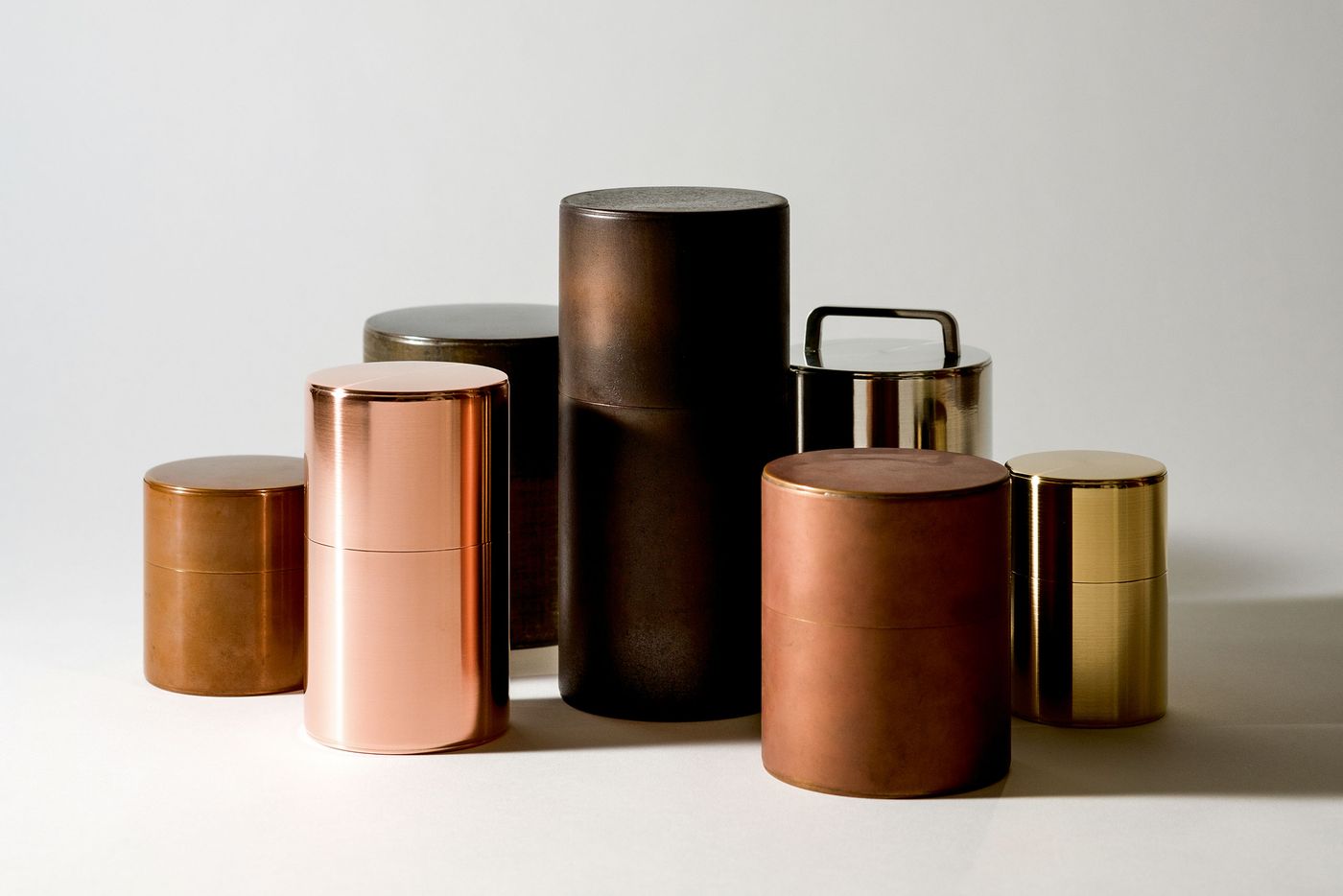 A collection of cylindrical metal containers in various heights and finishes, including copper, gold, and bronze, arranged artistically. A collection of cylindrical metal containers in various heights and finishes, including copper, gold, and bronze, arranged artistically.