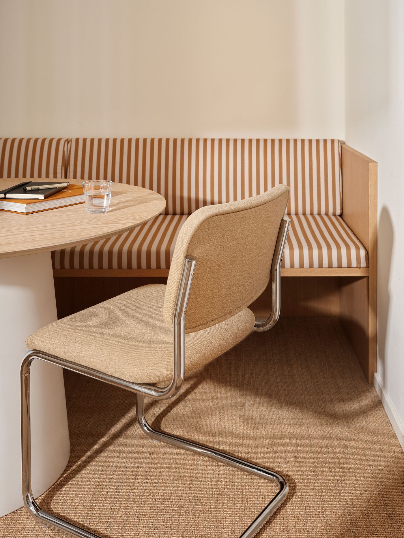 A cozy dining nook features a built-in bench with striped upholstery and a round, light wood table. A modern chair with a chrome frame and beige seat complements the natural textures and warm tones of the space.