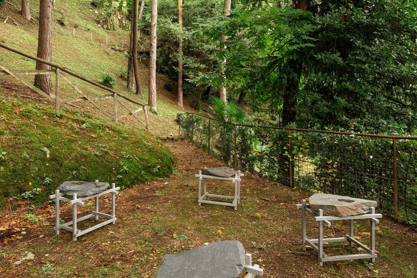 In a natural woodland setting, three contemporary stone sculptures are presented on minimalist metal stands. The stones, with their rough, natural textures, are juxtaposed against the metallic frames. The scene is nestled on a mossy, sloping hill dotted with trees and fallen leaves, with a rustic wooden fence and a subtle metal railing bordering the path. The placement emphasizes a Zen-like harmony between art and nature, highlighting organic forms.