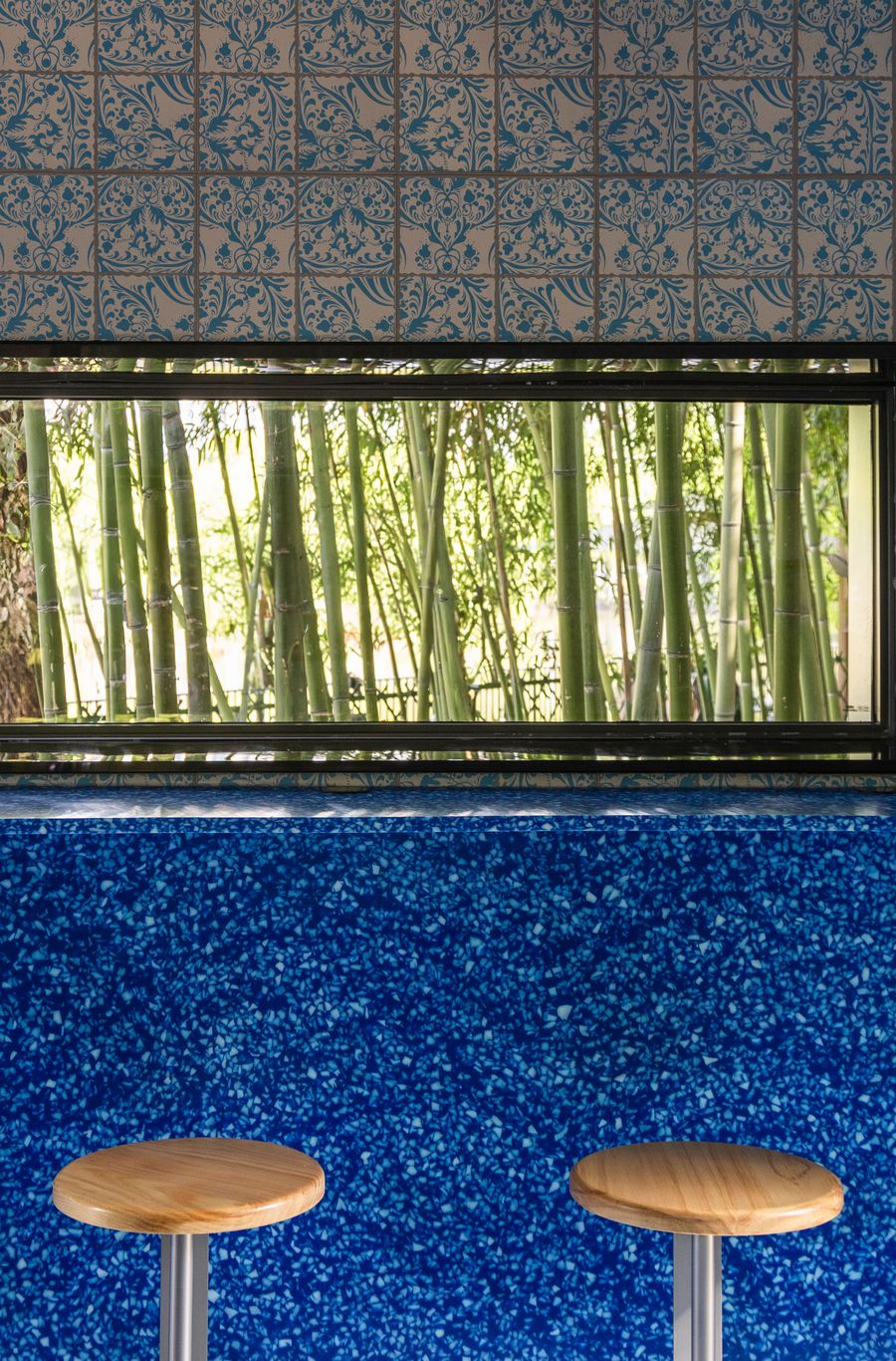 A vibrant and textured detail shot from Wutopia Lab's Lake House showcases a bold material palette. The lower half features a striking blue speckled surface, possibly terrazzo or a similar composite, supporting two minimalist wooden bar stools. Above, a framed horizontal window offers a natural view of a dense bamboo grove, creating a dynamic interplay between patterned tiles, modern materials, and biophilic elements. A vibrant and textured detail shot from Wutopia Lab's Lake House showcases a bold material palette. The lower half features a striking blue speckled surface, possibly terrazzo or a similar composite, supporting two minimalist wooden bar stools. Above, a framed horizontal window offers a natural view of a dense bamboo grove, creating a dynamic interplay between patterned tiles, modern materials, and biophilic elements.