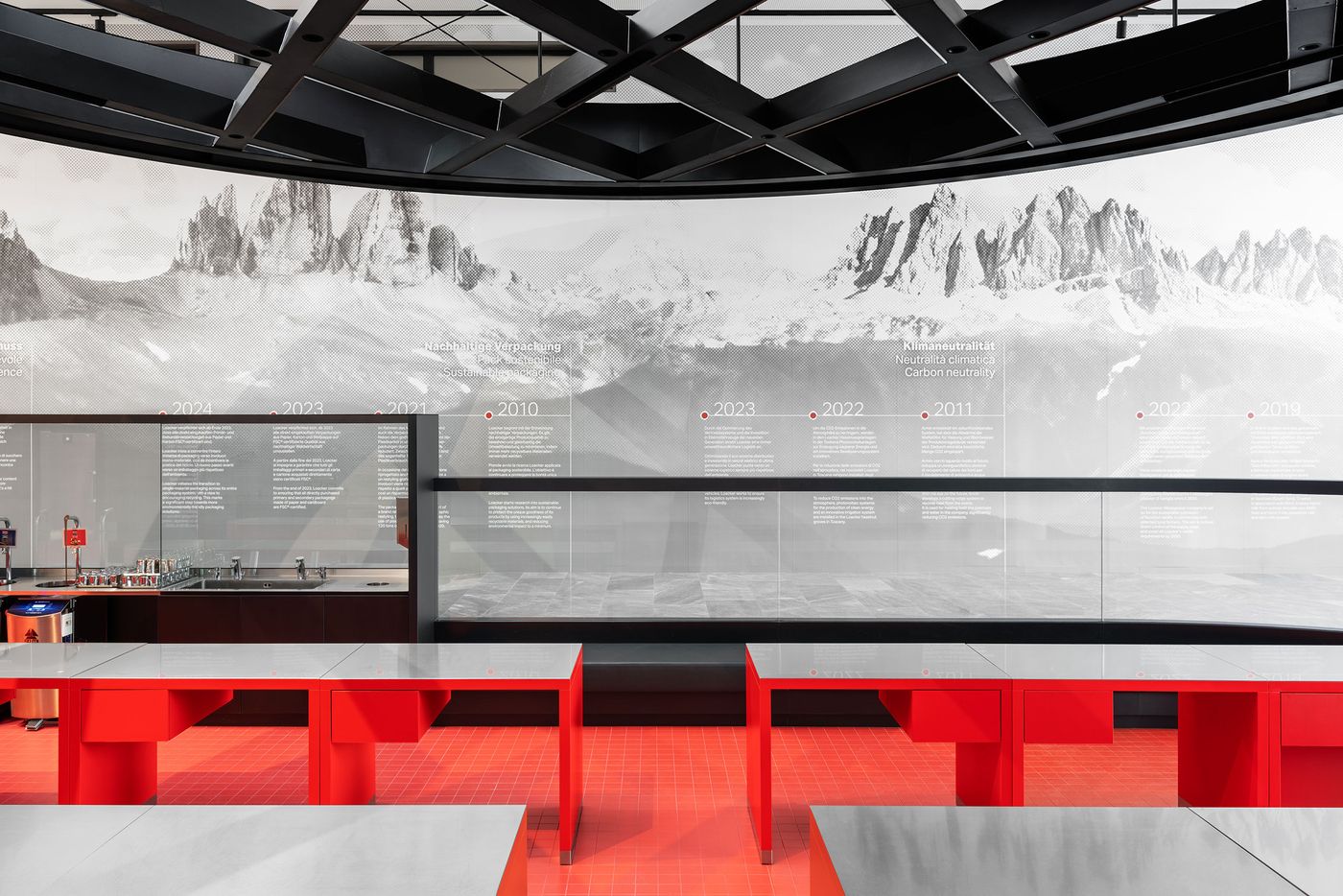 An amphitheater-style presentation space descends toward panoramic mountain imagery, with bold red seating creating dramatic focal point. The curved ceiling treatment and systematic lighting demonstrate how exhibition design can transform corporate storytelling into immersive spatial experience through careful choreography of view and movement. An amphitheater-style presentation space descends toward panoramic mountain imagery, with bold red seating creating dramatic focal point. The curved ceiling treatment and systematic lighting demonstrate how exhibition design can transform corporate storytelling into immersive spatial experience through careful choreography of view and movement.