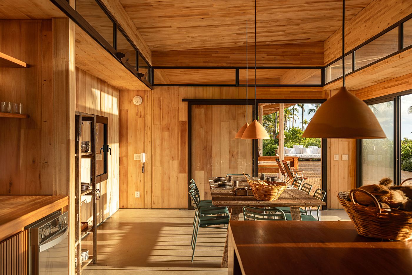 A modern kitchen and dining space combines sleek wooden surfaces with industrial-style shelving and ceramic pendant lights. Large sliding doors offer glimpses of an outdoor lounging area, blending functional elegance with a relaxed, open-plan living ideal for a tropical setting. A modern kitchen and dining space combines sleek wooden surfaces with industrial-style shelving and ceramic pendant lights. Large sliding doors offer glimpses of an outdoor lounging area, blending functional elegance with a relaxed, open-plan living ideal for a tropical setting.