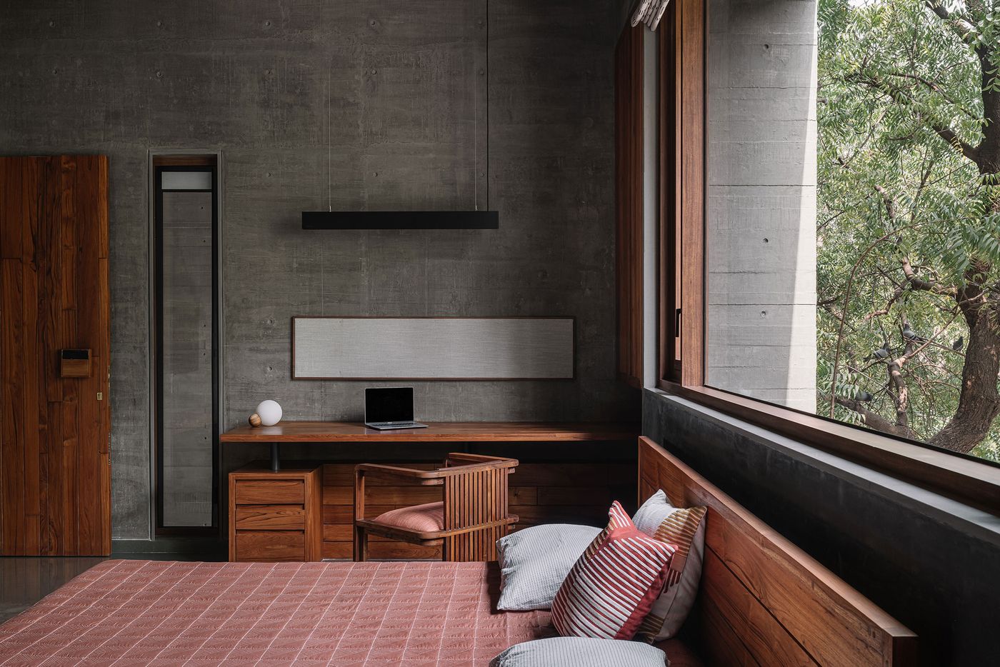 A minimalist bedroom featuring a bespoke wooden bed frame and an integrated desk area along a concrete wall. A suspended black linear light fixture illuminates the workspace, while a large horizontal window offers expansive views of lush outdoor greenery. The raw concrete and warm wood create a balanced, earthy aesthetic.
