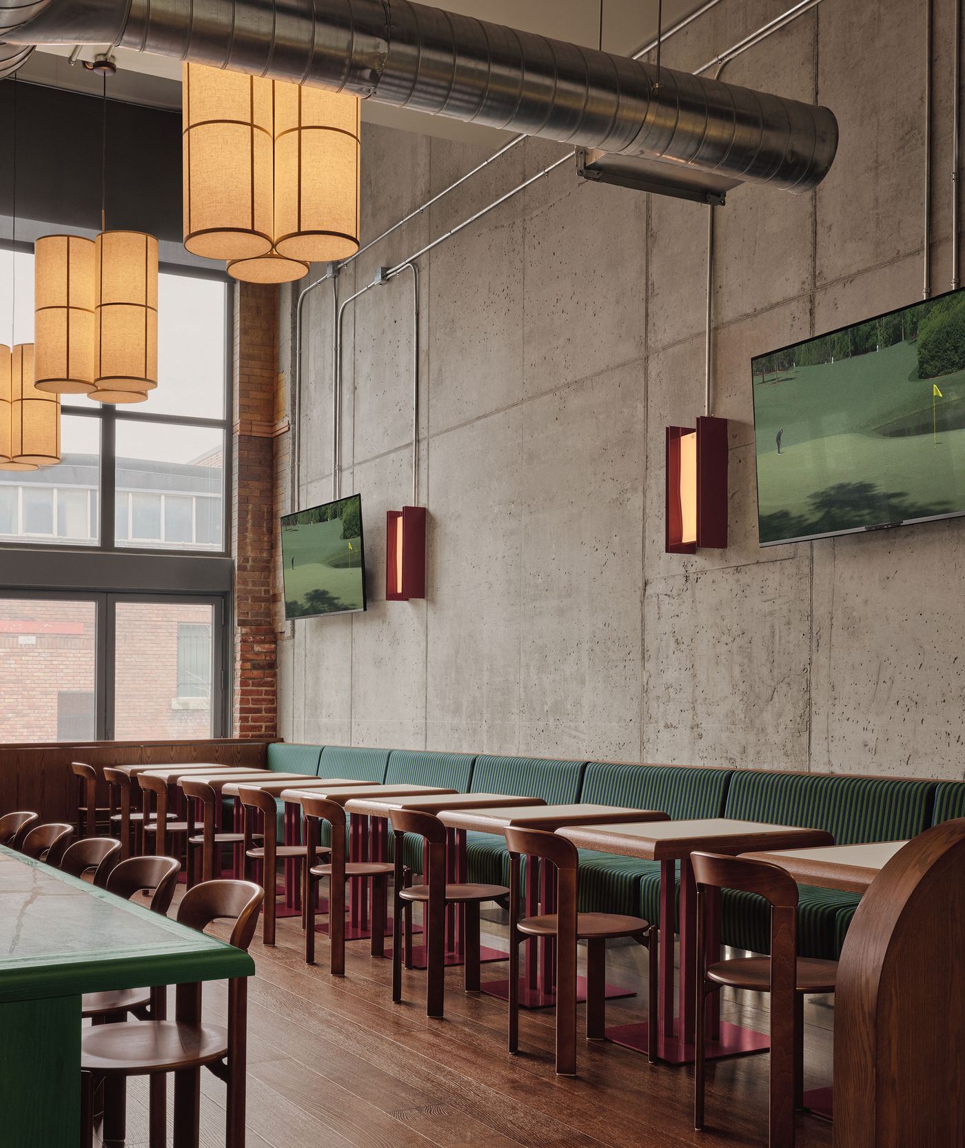 A modern restaurant interior featuring wooden tables, green seating, large windows, and decorative pendant lights. Two TVs display golf. A modern restaurant interior featuring wooden tables, green seating, large windows, and decorative pendant lights. Two TVs display golf.