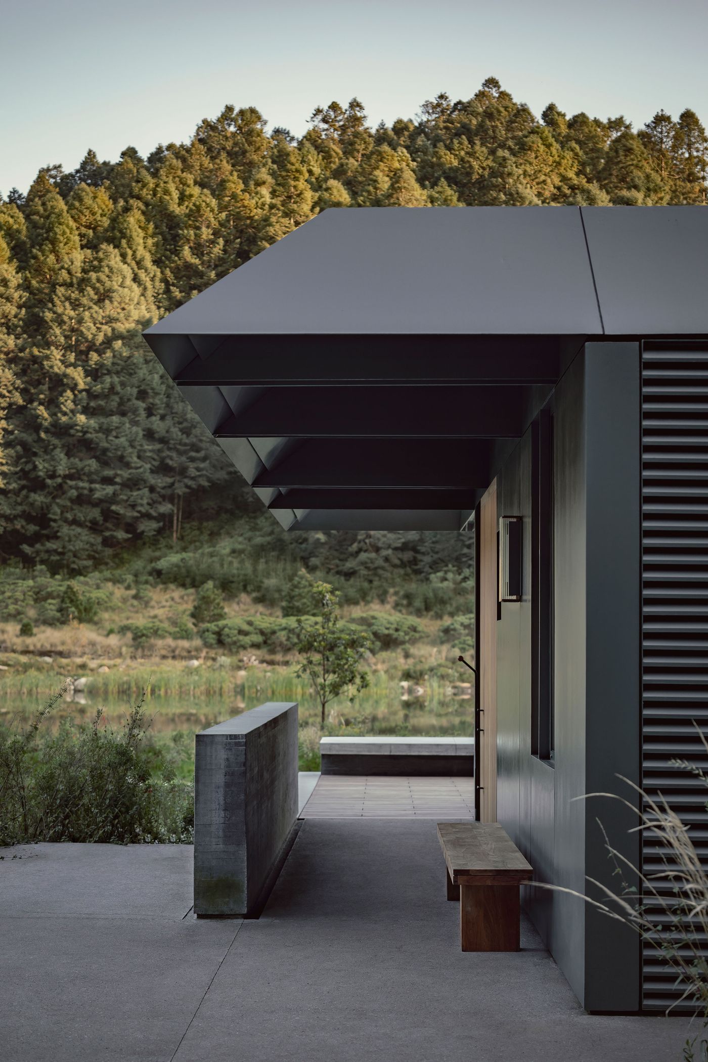 Modern house exterior with a slanted roof, concrete bench, and lush forest backdrop reflecting in a tranquil water body.