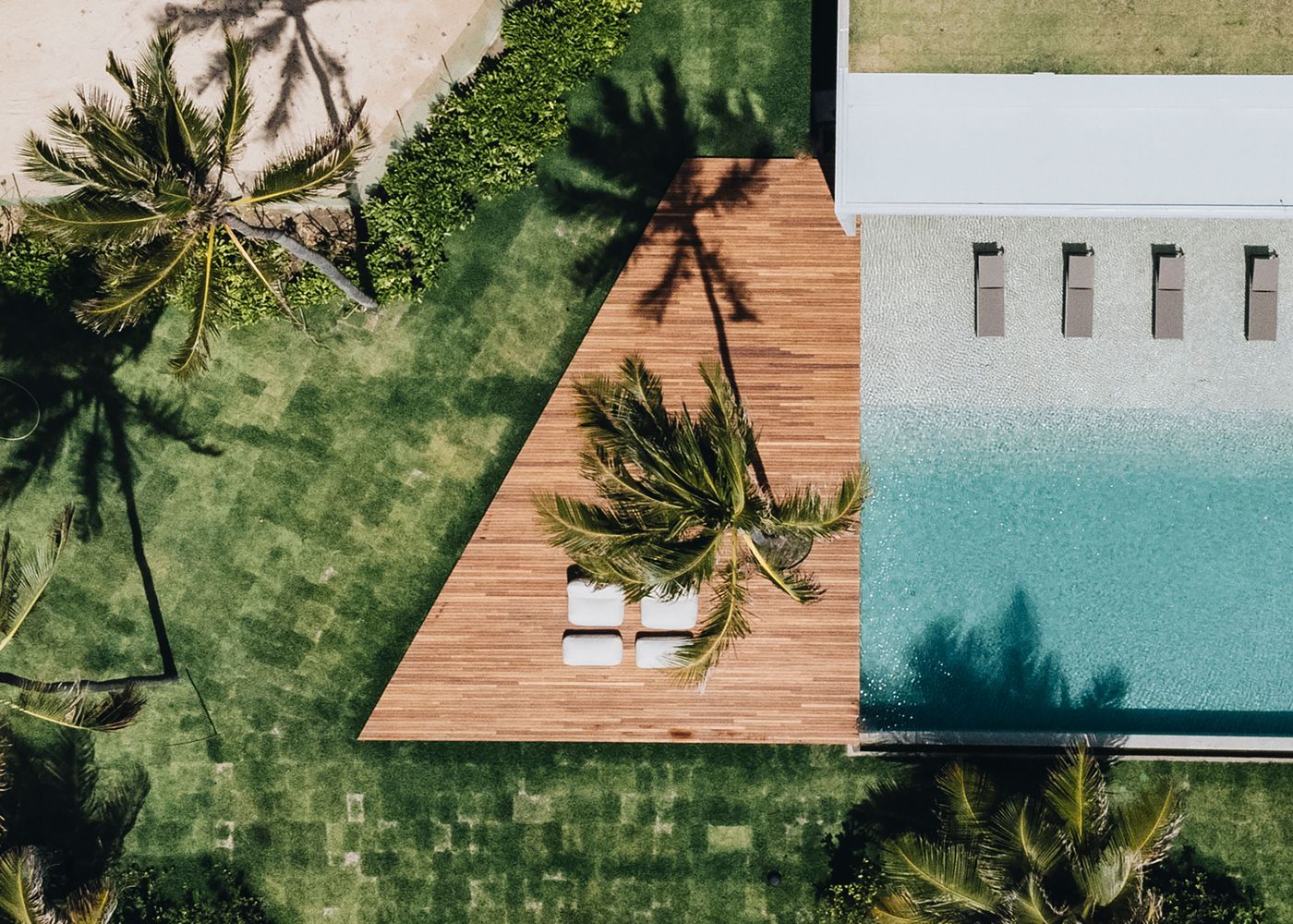 Aerial view of a lush green garden featuring a wooden deck with lounge chairs by a sleek, turquoise infinity pool.
