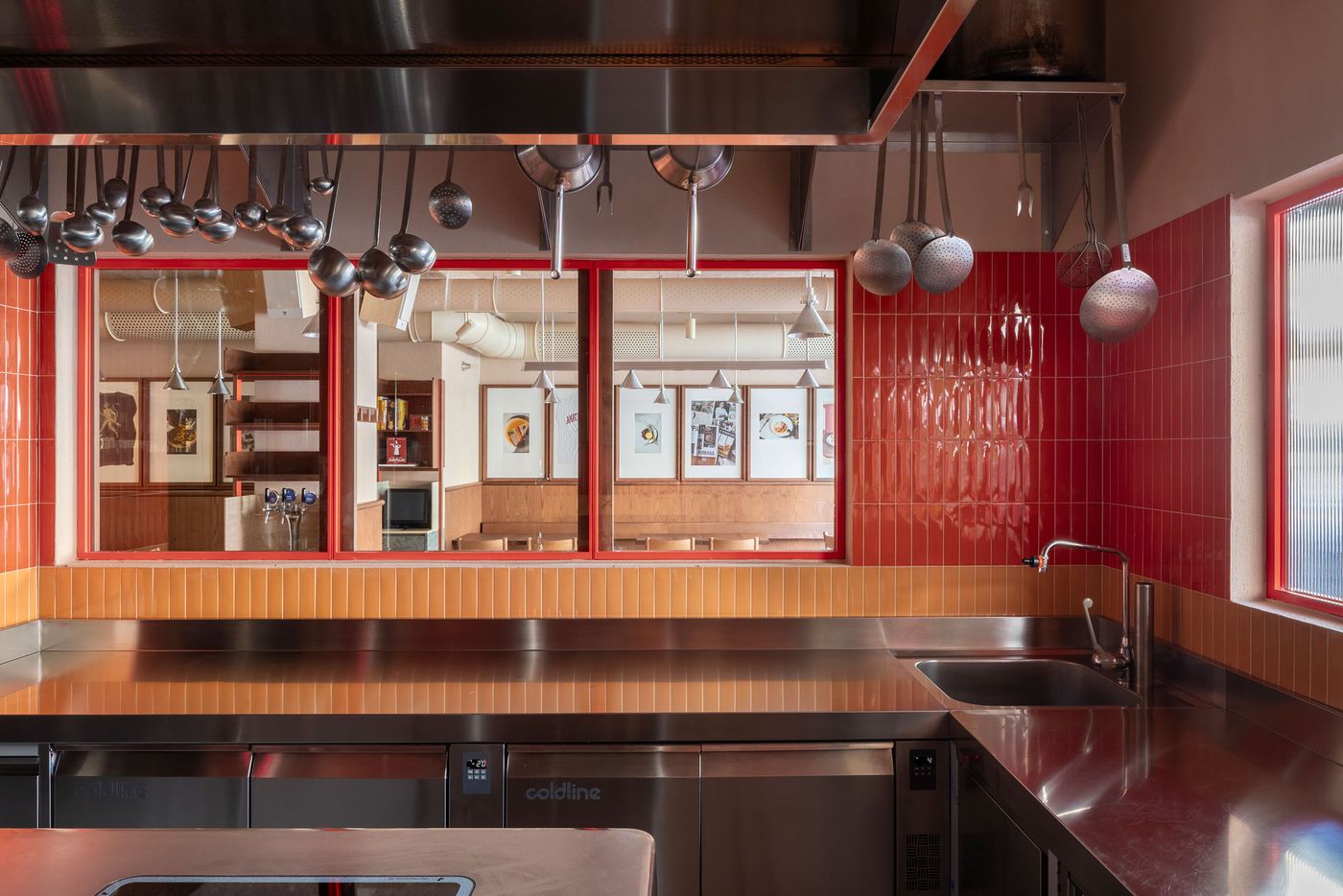 A dynamic glimpse into Santo Palato's kitchen pass, designed by Naessi Studio, showcasing a blend of functionality and vibrant color. Polished stainless steel countertops contrast with a striking red- and orange-tiled backsplash. Red-framed windows offer a peek into the dining area, while hanging kitchen utensils create an industrial yet inviting aesthetic. A dynamic glimpse into Santo Palato's kitchen pass, designed by Naessi Studio, showcasing a blend of functionality and vibrant color. Polished stainless steel countertops contrast with a striking red- and orange-tiled backsplash. Red-framed windows offer a peek into the dining area, while hanging kitchen utensils create an industrial yet inviting aesthetic.